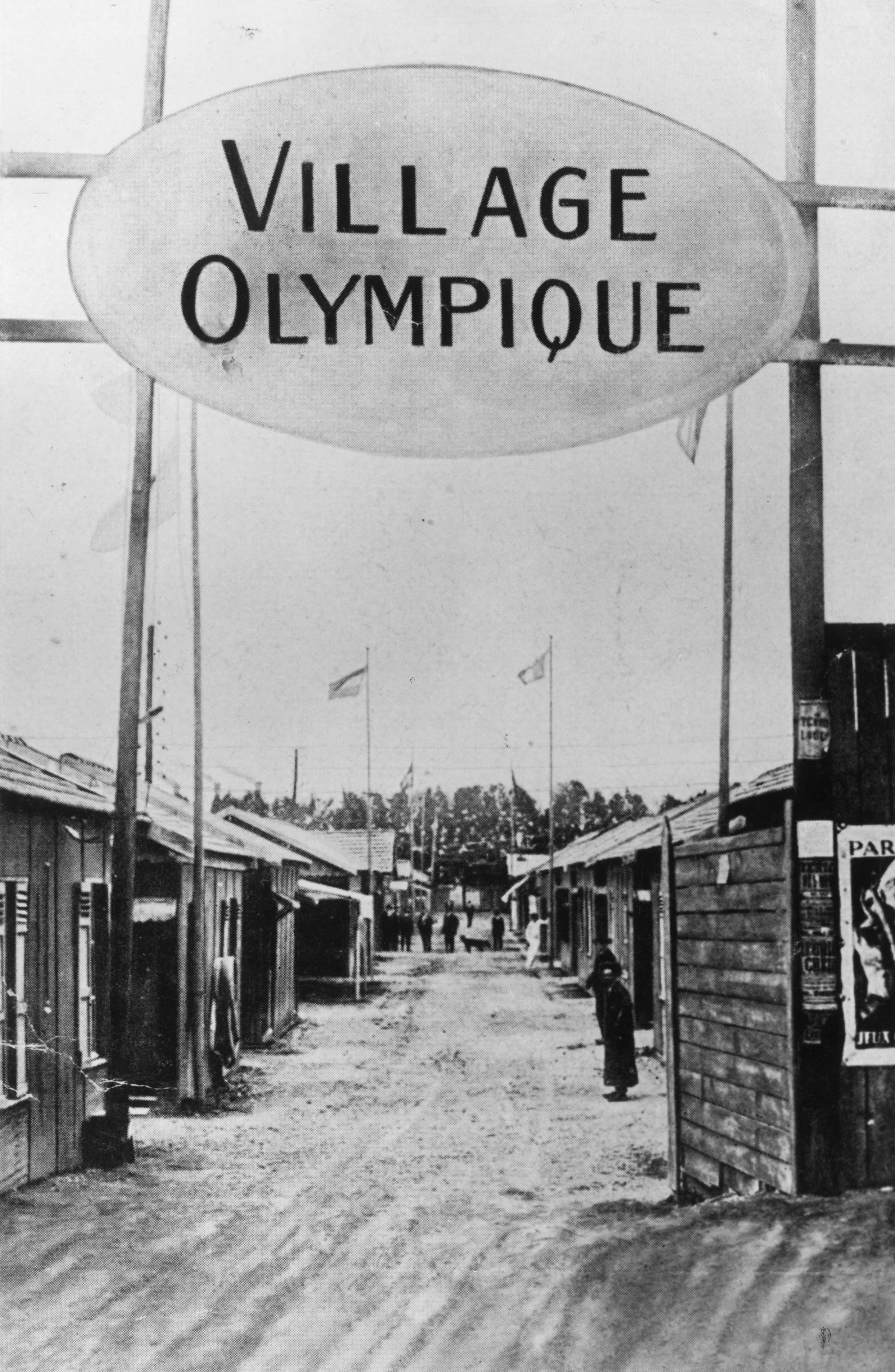 Historic photograph of the Olympic Village with small wooden buildings and a large overhead sign reading &quot;Village Olympique&quot;