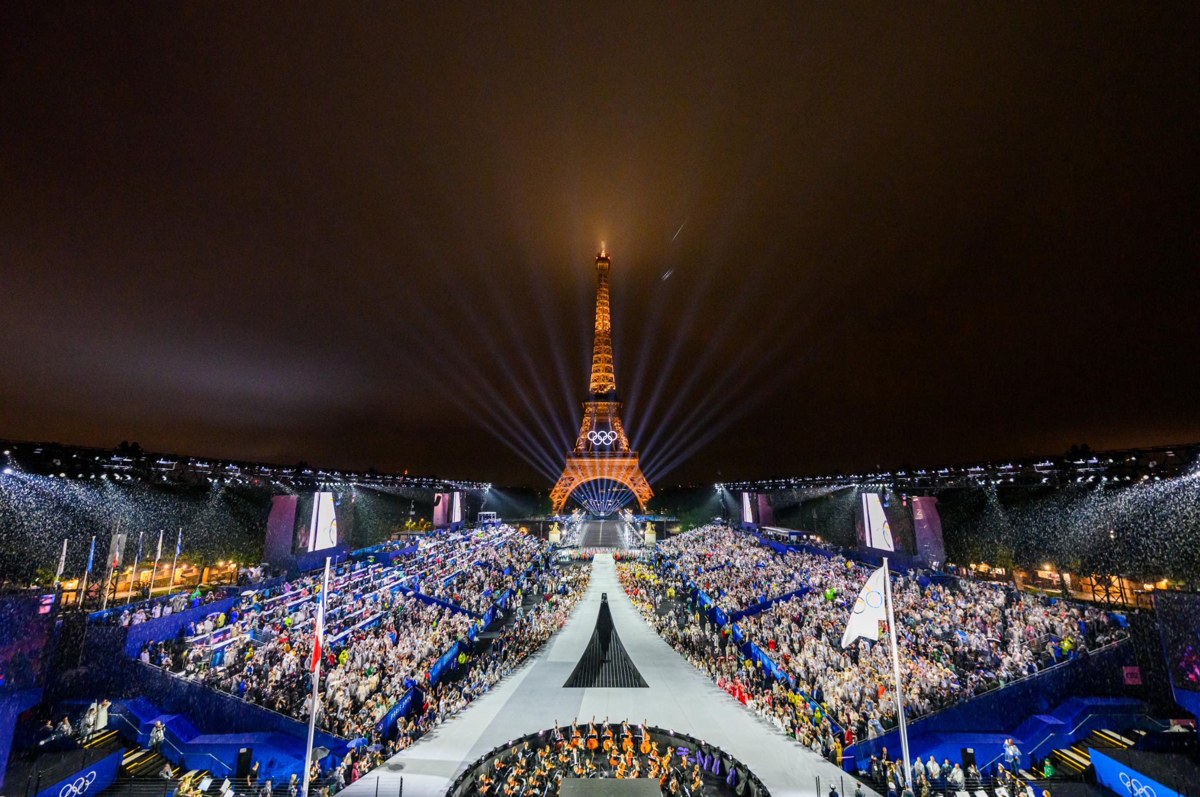 Large crowd at a nighttime event in front of the illuminated Eiffel Tower, with light beams and fireworks, celebrating Paris 2024 Olympics