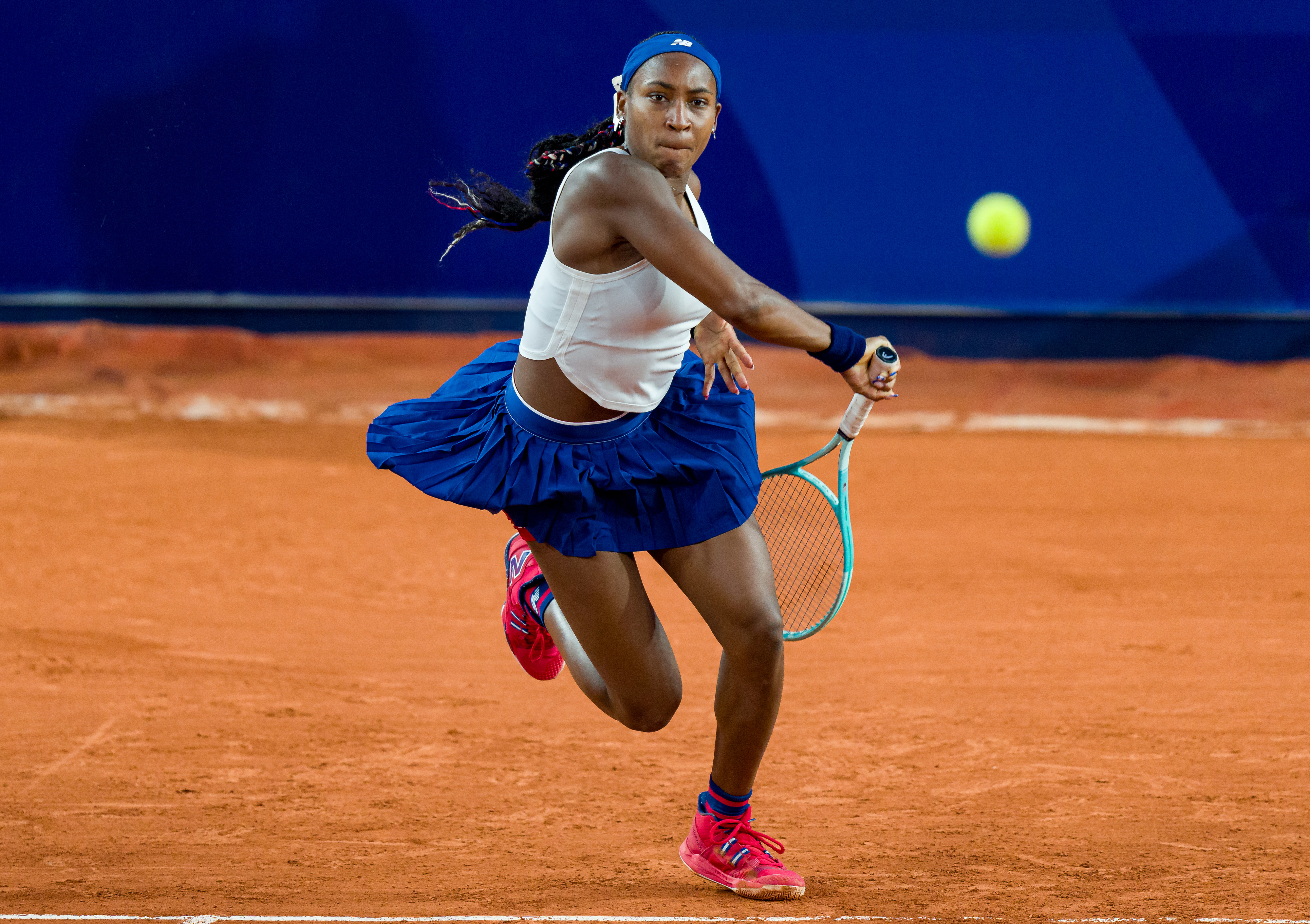 Coco Gauff in action on a tennis court, wearing a sleeveless top, a skirt, a headband, and wristbands, preparing to hit a tennis ball with her racquet