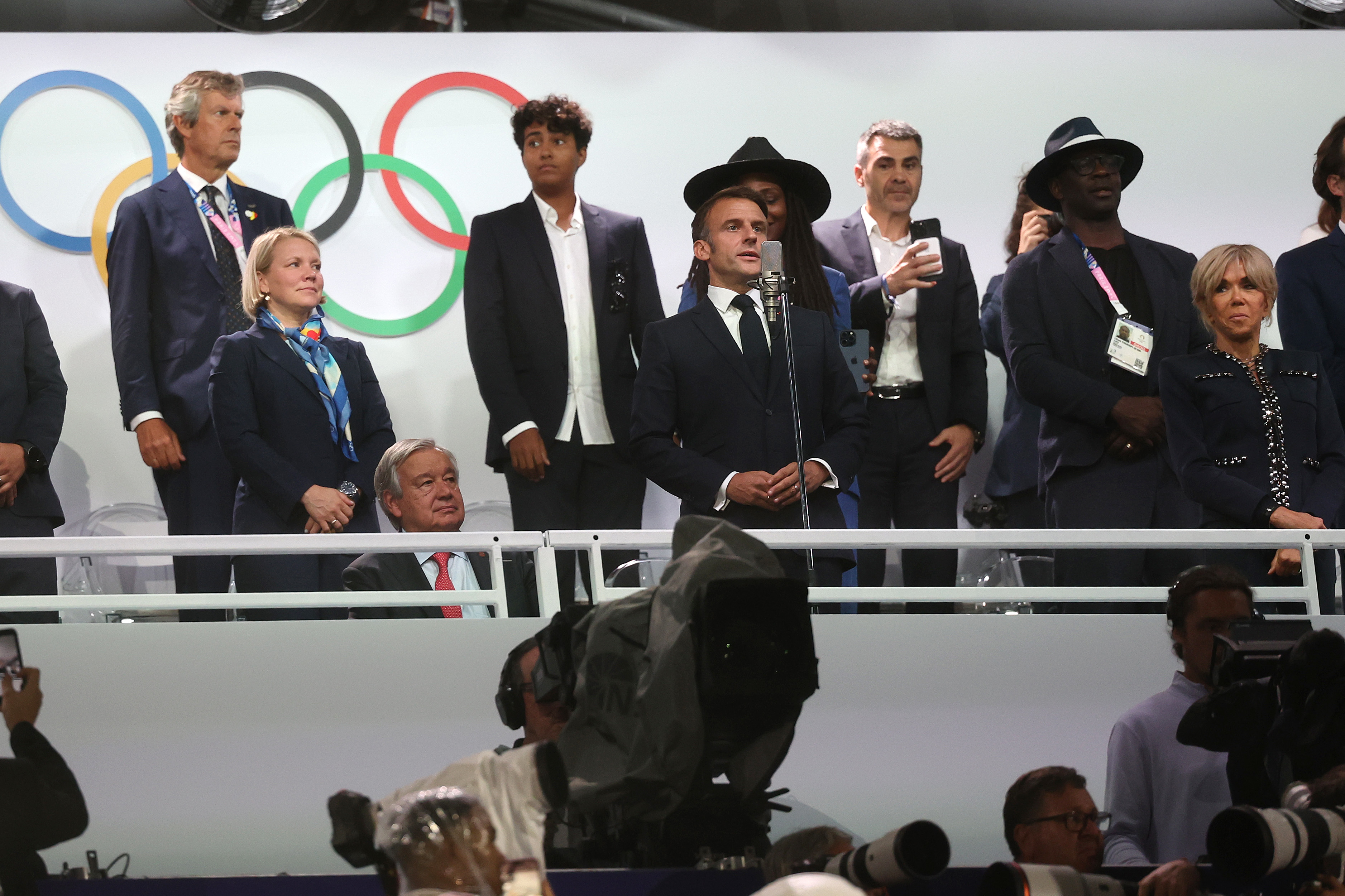 Emmanuel Macron, António Guterres, and other officials stand in front of the Olympic rings at a sports event. Macron is speaking at a podium