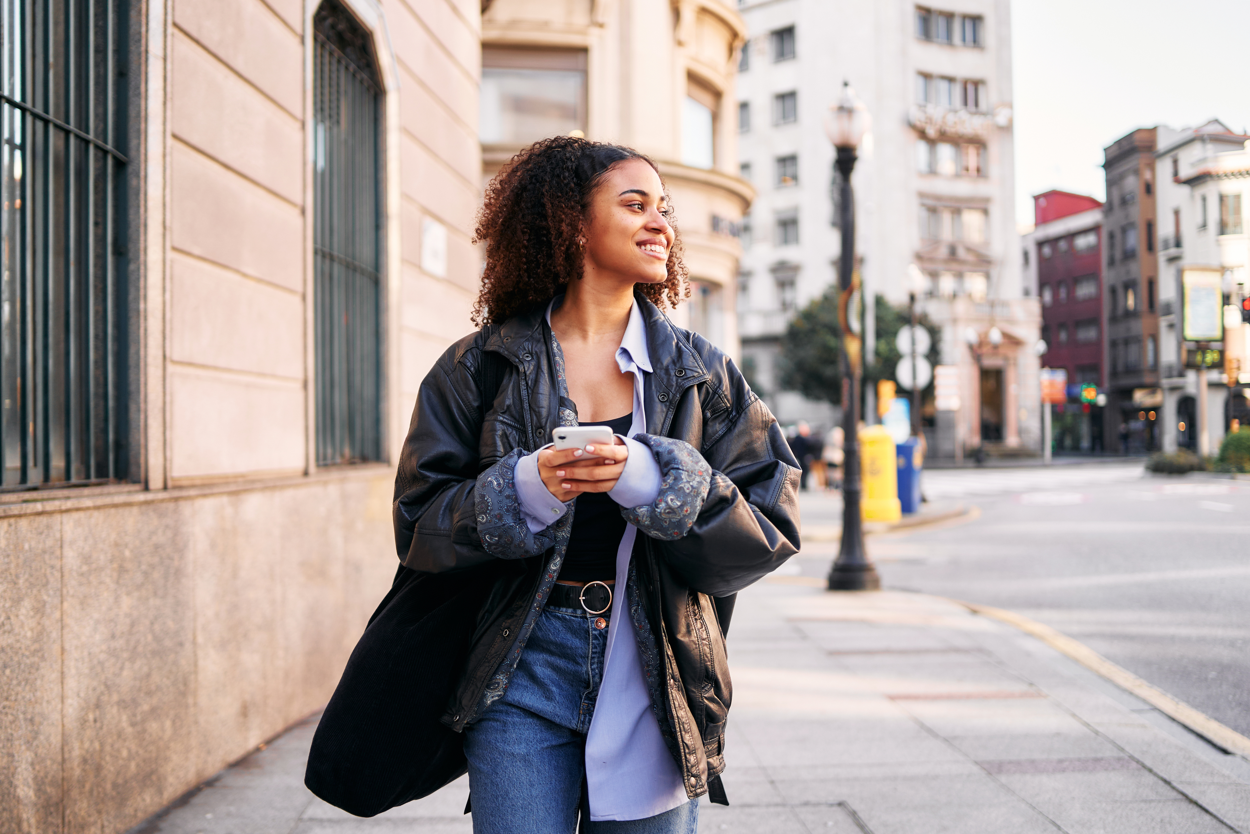 A woman stands on a city street, smiling while holding a phone and wearing a large jacket over a button-down shirt and jeans