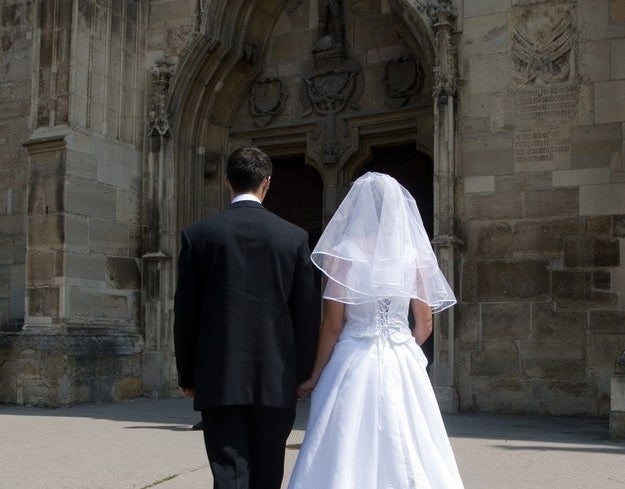 A bride and groom, both in formal wedding attire, walk hand-in-hand towards a large, ornate church entrance. Their backs are facing the camera