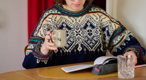 Person reading a book at a table while holding a mug. Wearing a patterned sweater