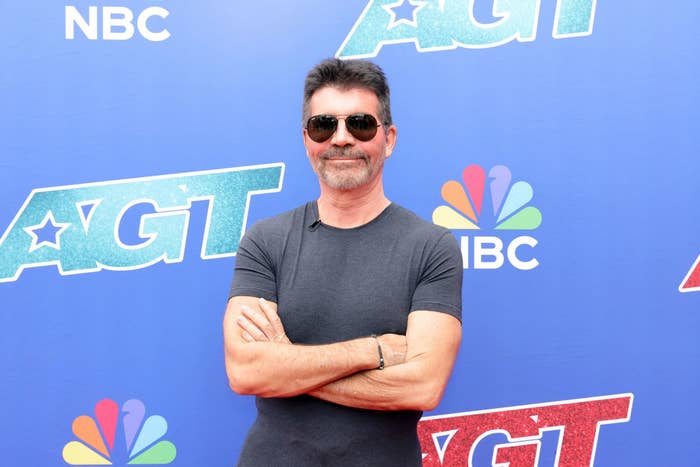 Simon Cowell stands on a red carpet in front of an &quot;America&#x27;s Got Talent&quot; backdrop, wearing a fitted short-sleeve shirt and sunglasses, smiling with arms crossed