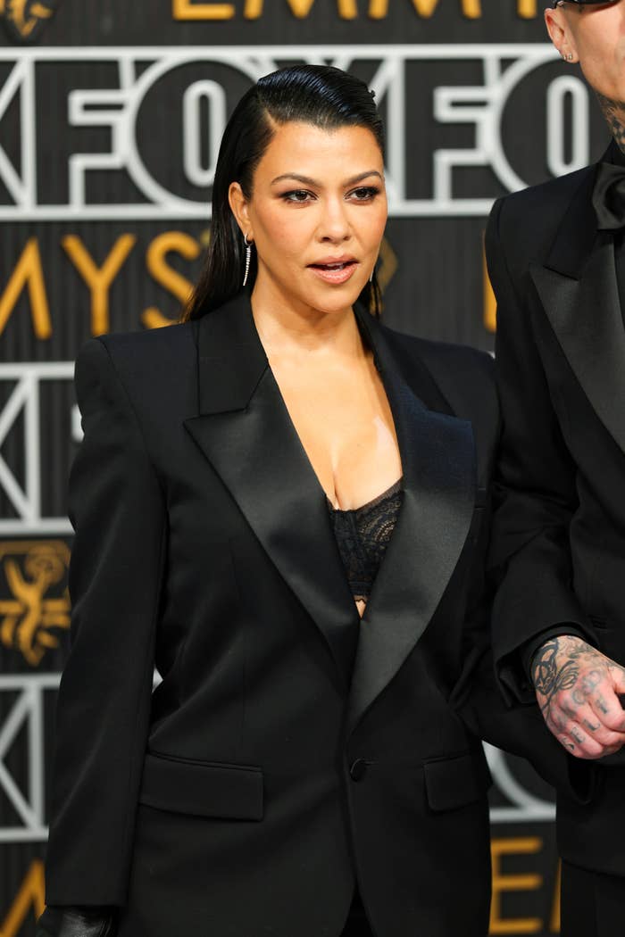 Kourtney Kardashian on a red carpet, wearing a sleek dark blazer over a low-cut top