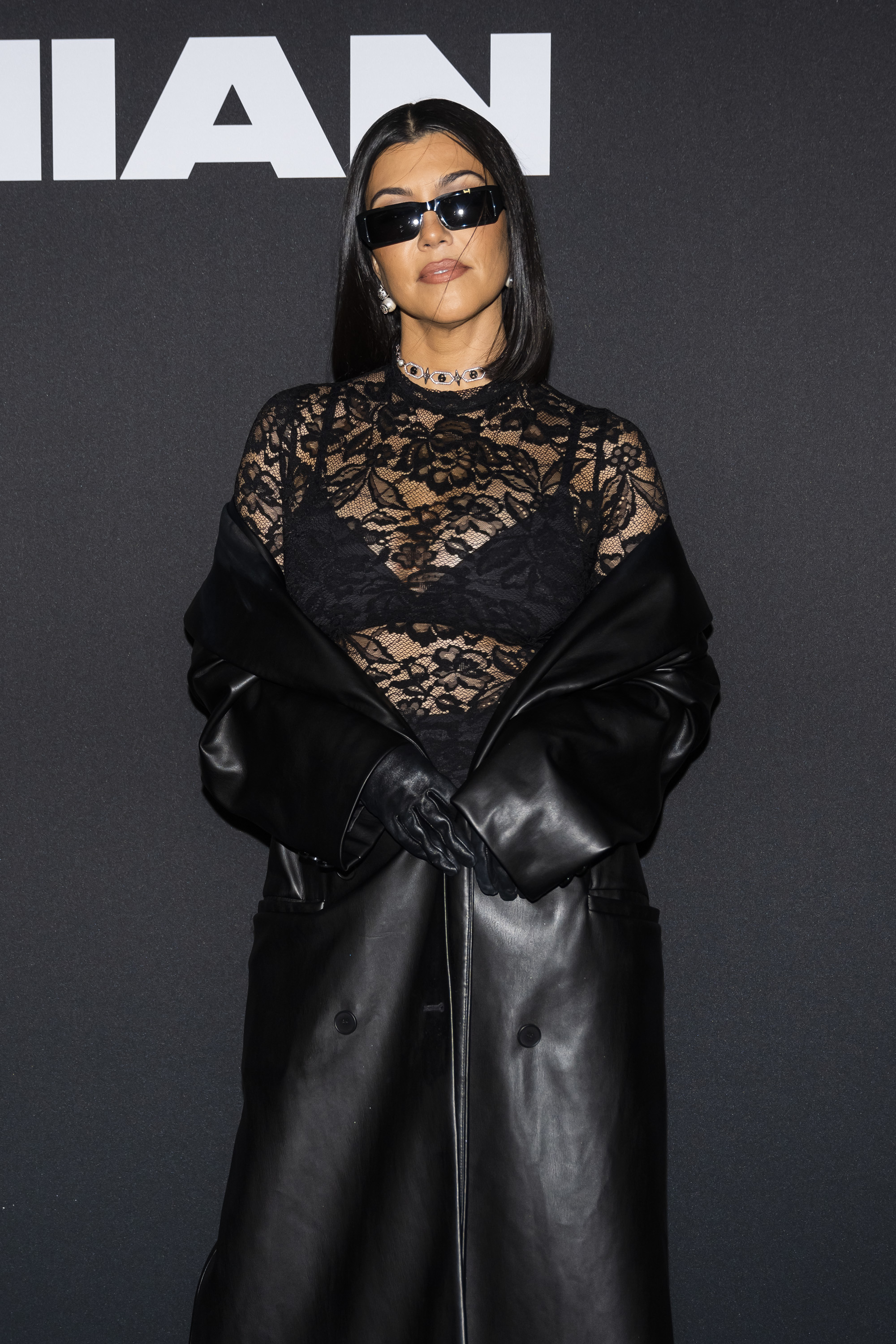 Kourtney Kardashian at an event in a lace top with a bra underneath, leather gloves, and a long leather coat, posing against a dark background