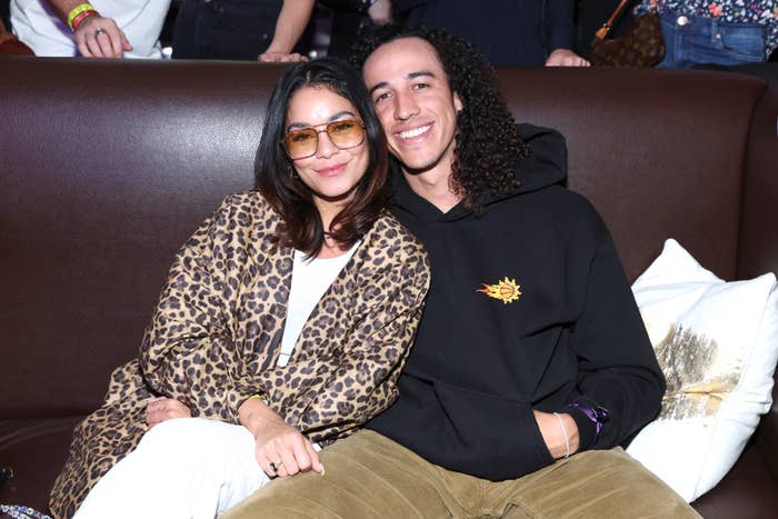Vanessa Hudgens, in a leopard print jacket, and Cole Tucker, in a casual hoodie, sit together on a couch, smiling at the camera
