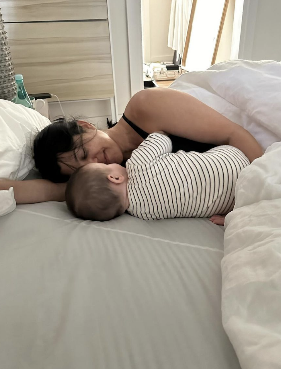 Kourtney Kardashian lies in bed facing her baby in striped clothing, nose to nose, while both rest on their sides under a comforter