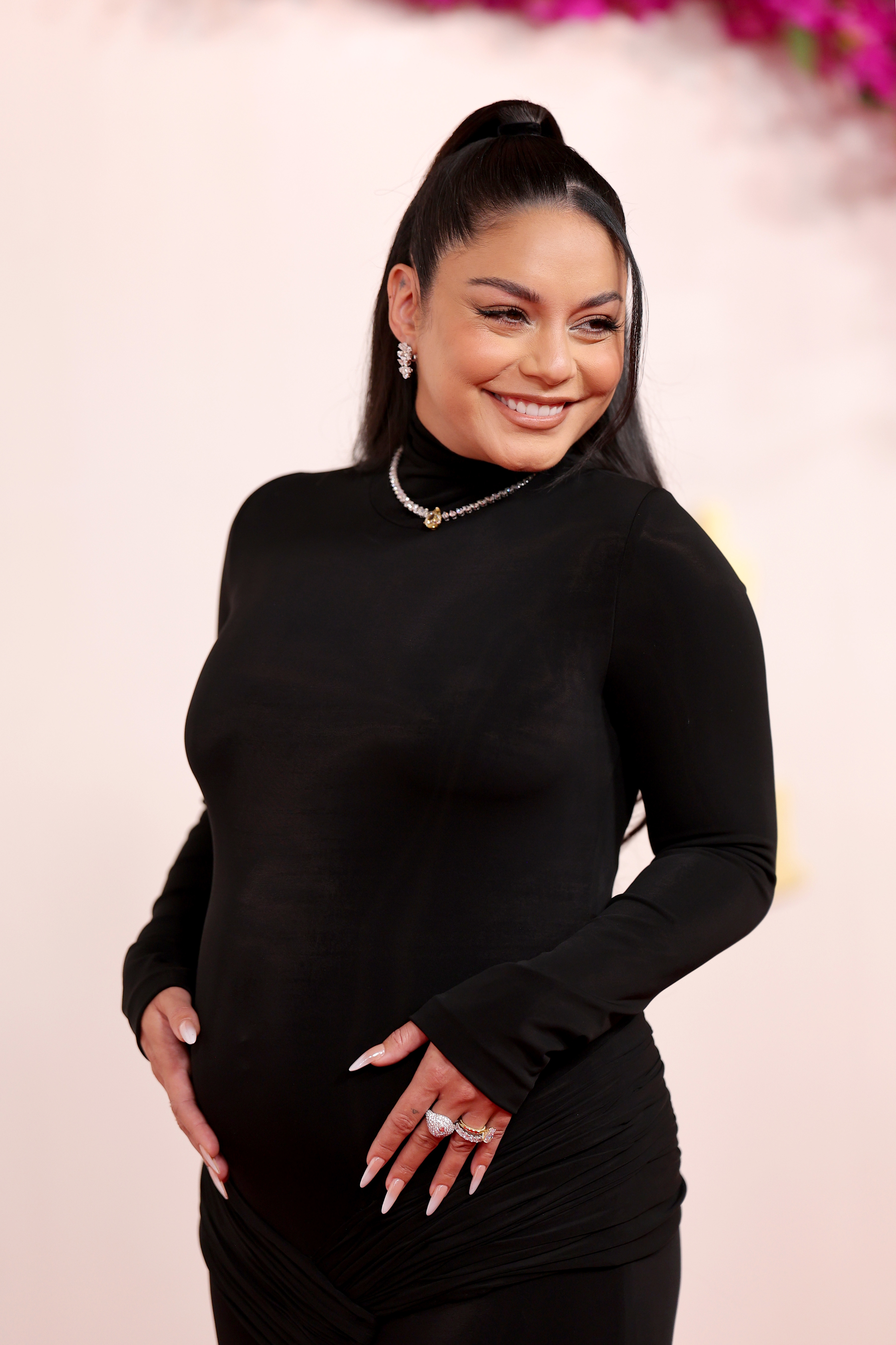 Vanessa Hudgens cradles her baby bump, smiling in a fitted black gown with long sleeves and a high neckline on the red carpet