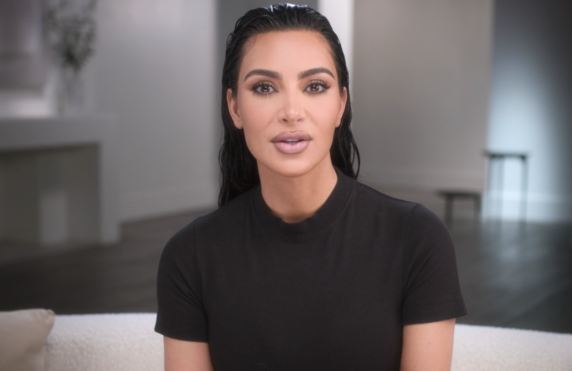 Kim Kardashian, with wet hair and wearing a simple shirt, sits on a light-colored couch in a modern, minimalistic room