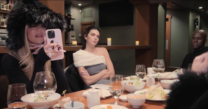 Khloé Kardashian FaceTime&#x27;swhile Kendall Jenner, in a stylish off-shoulder dress, and others sit at a dinner table with various dishes