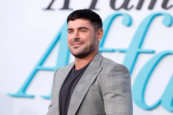 Zac Efron stands smiling at an event, wearing a checkered blazer over a dark shirt, with a &quot;A Family Affair&quot; sign in the background