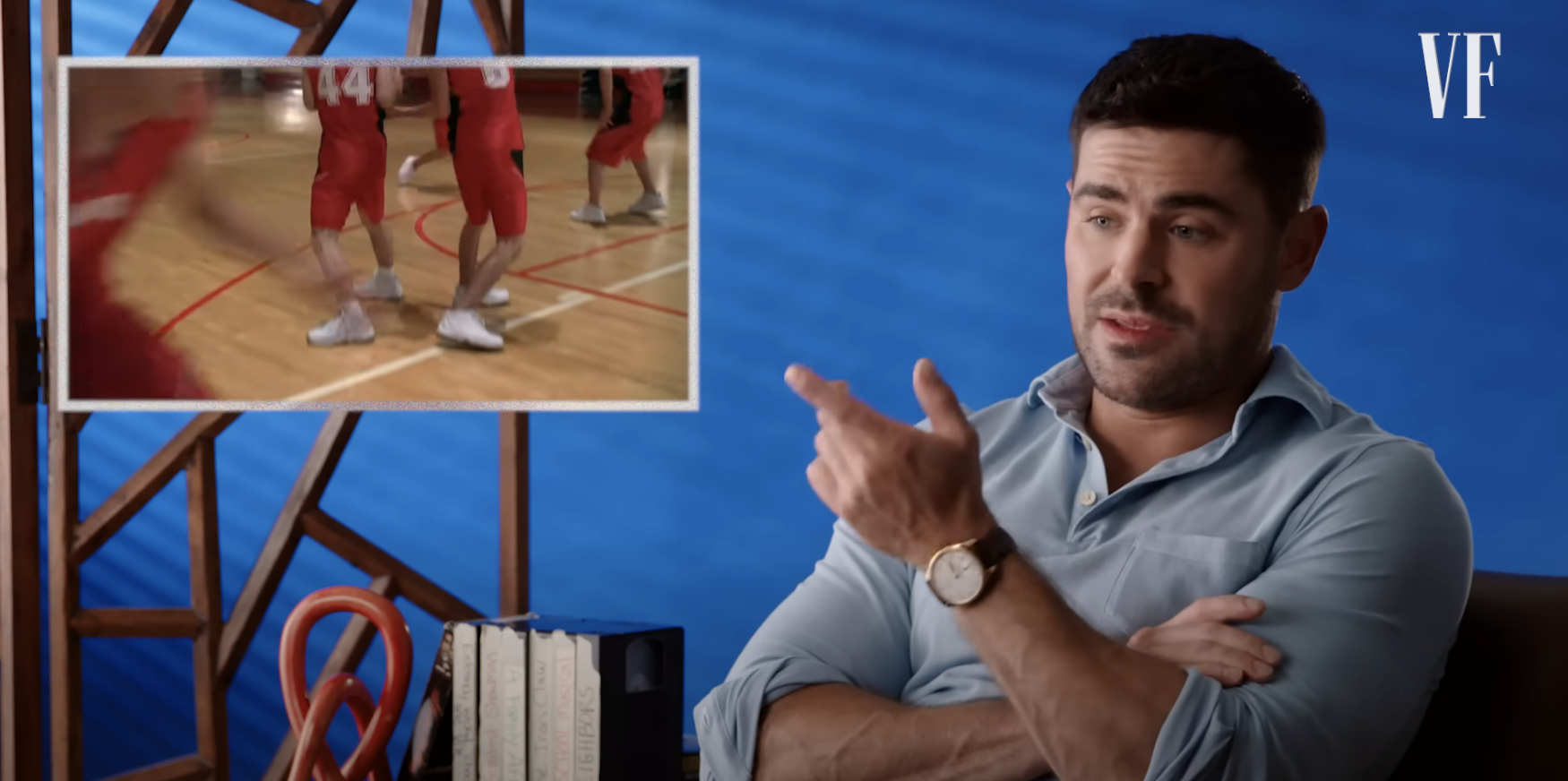 Zac Efron discusses a scene from a basketball game shown in an inset. He&#x27;s seated, wearing a light shirt, pointing to the inset image. Vanity Fair logo present