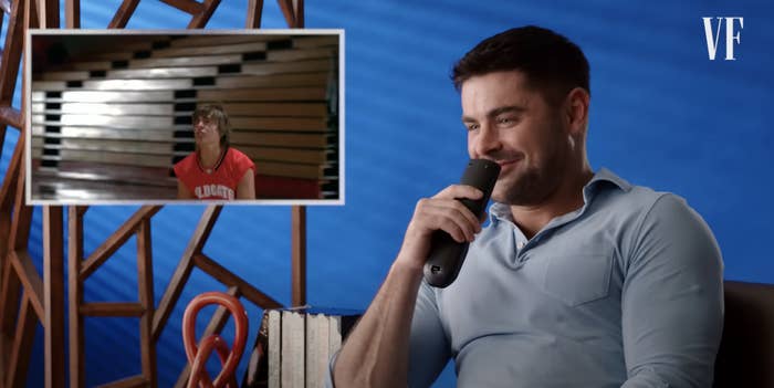 Zac Efron is being interviewed and shown an old photo of himself in a red jersey, holding a microphone, with books and a wood-patterned background