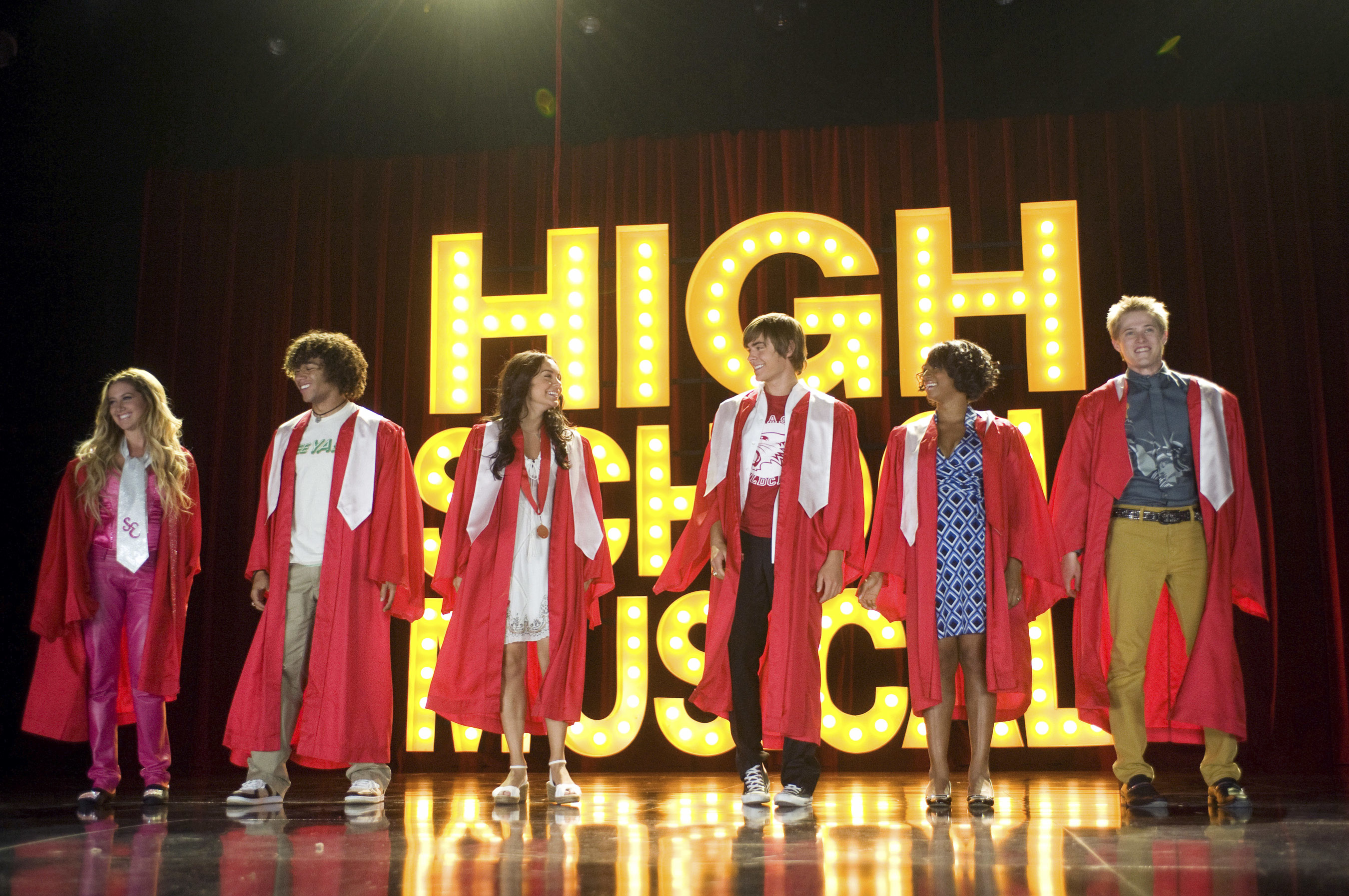 Ashley Tisdale, Corbin Bleu, Vanessa Hudgens, Zac Efron, Monique Coleman, and Lucas Grabeel stand in red graduation robes on stage in front of &quot;High School Musical&quot; sign