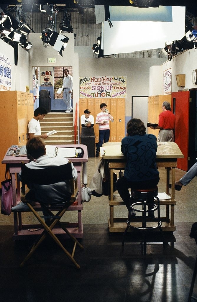 Actors rehearsing a scene on a high school hallway set for a TV show. Production crew members are observing and managing the scene