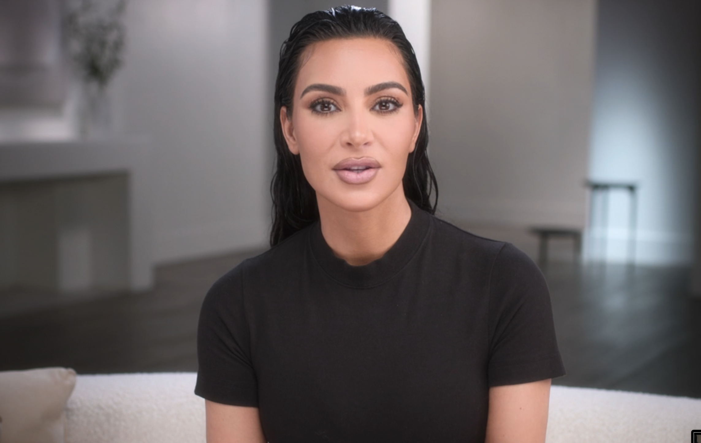 Kim Kardashian sits on a couch in a minimalistic room, wearing a simple black top, with slicked-back hair and natural makeup
