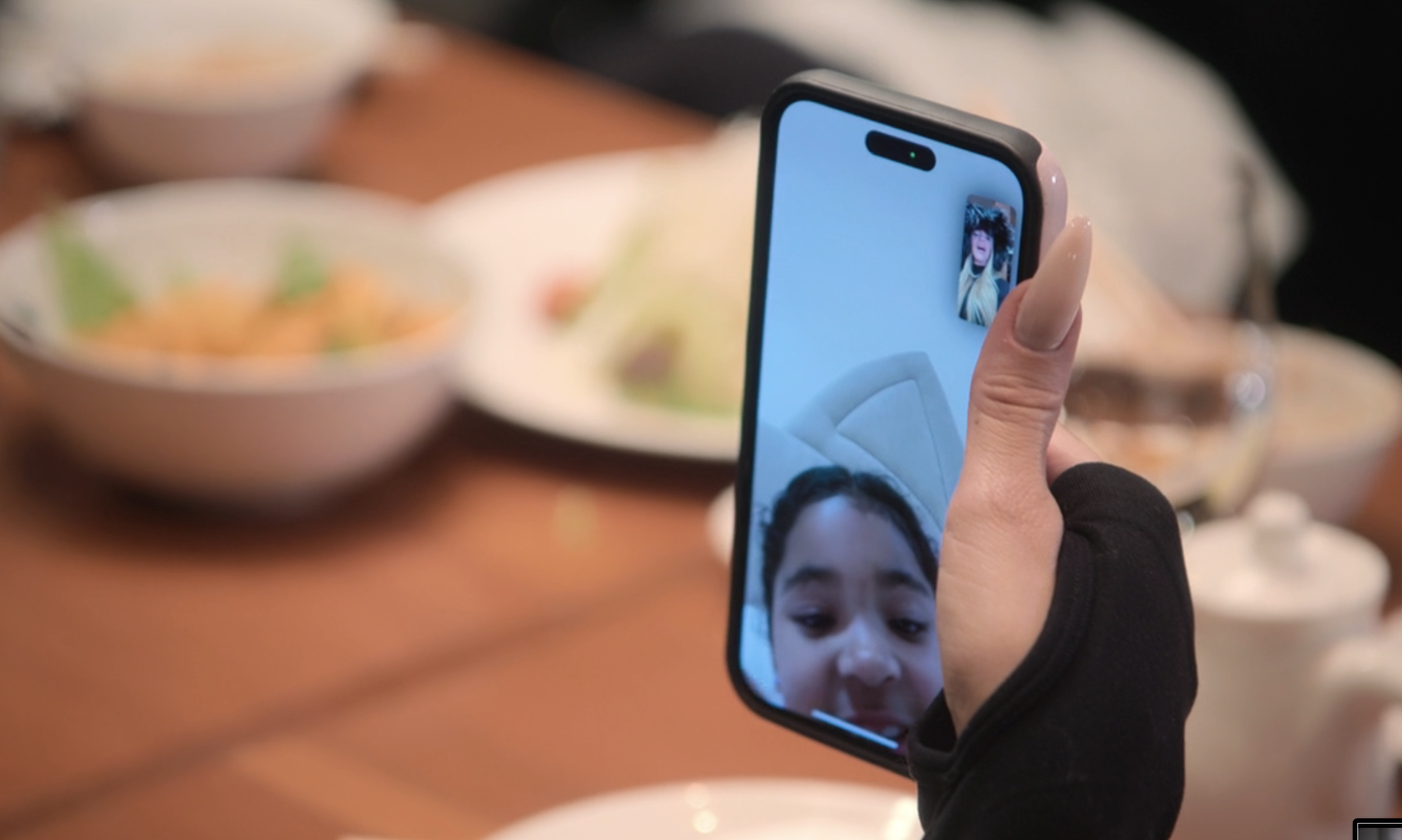 Close-up of a hand holding a phone showing a video call with two people. In the background, a dining table with several plates of food is visible