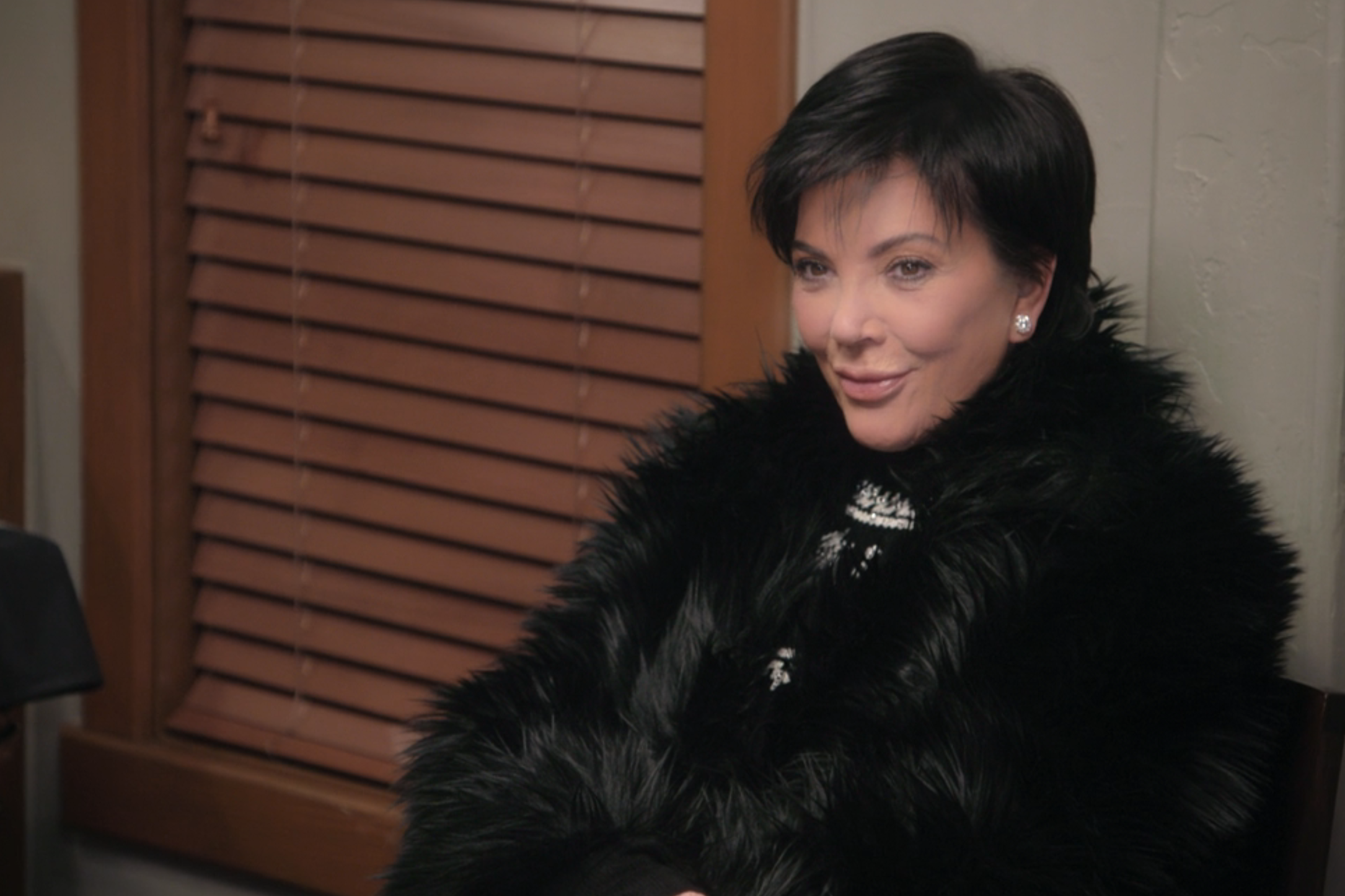 Kris Jenner is seated indoors, wearing a black fur coat with a patterned top underneath. She is smiling and looking slightly off-camera
