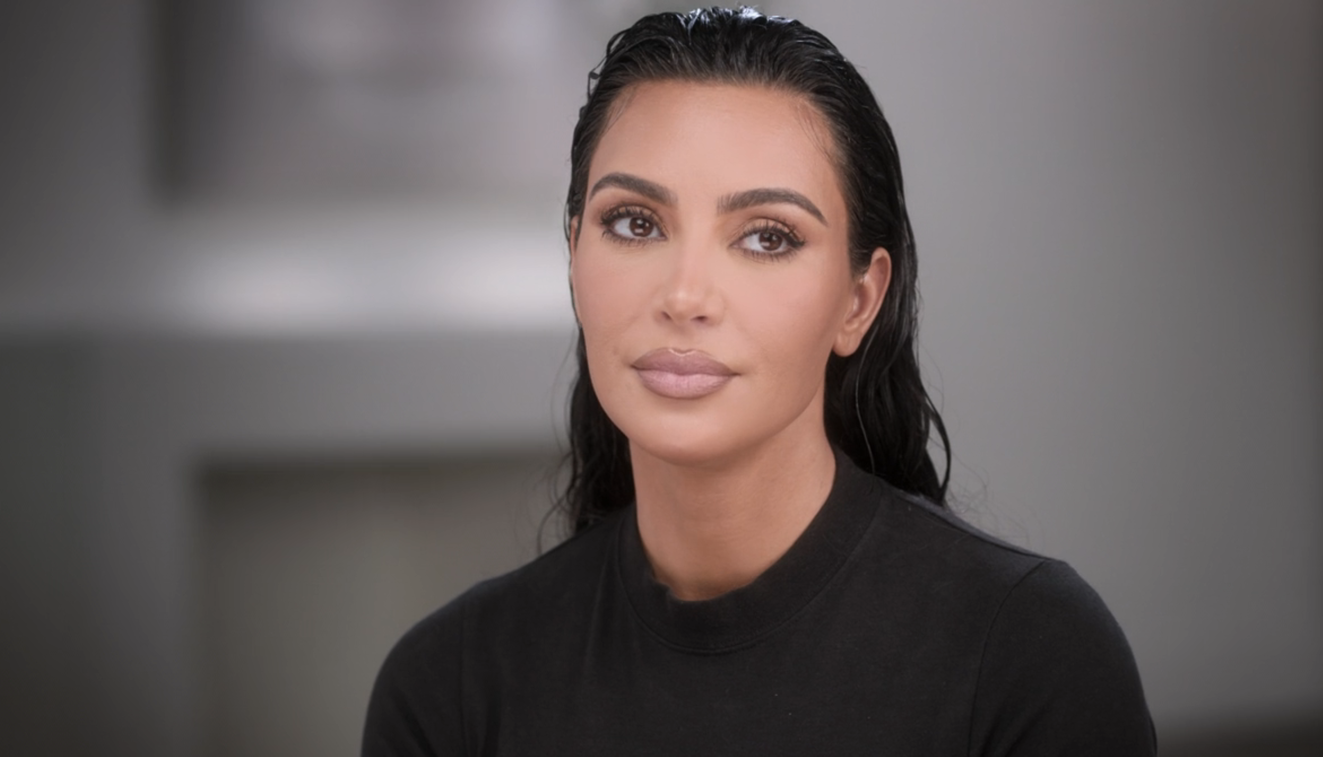 Kim Kardashian, hair slicked back, wearing a fitted black top, appearing in an interview setting