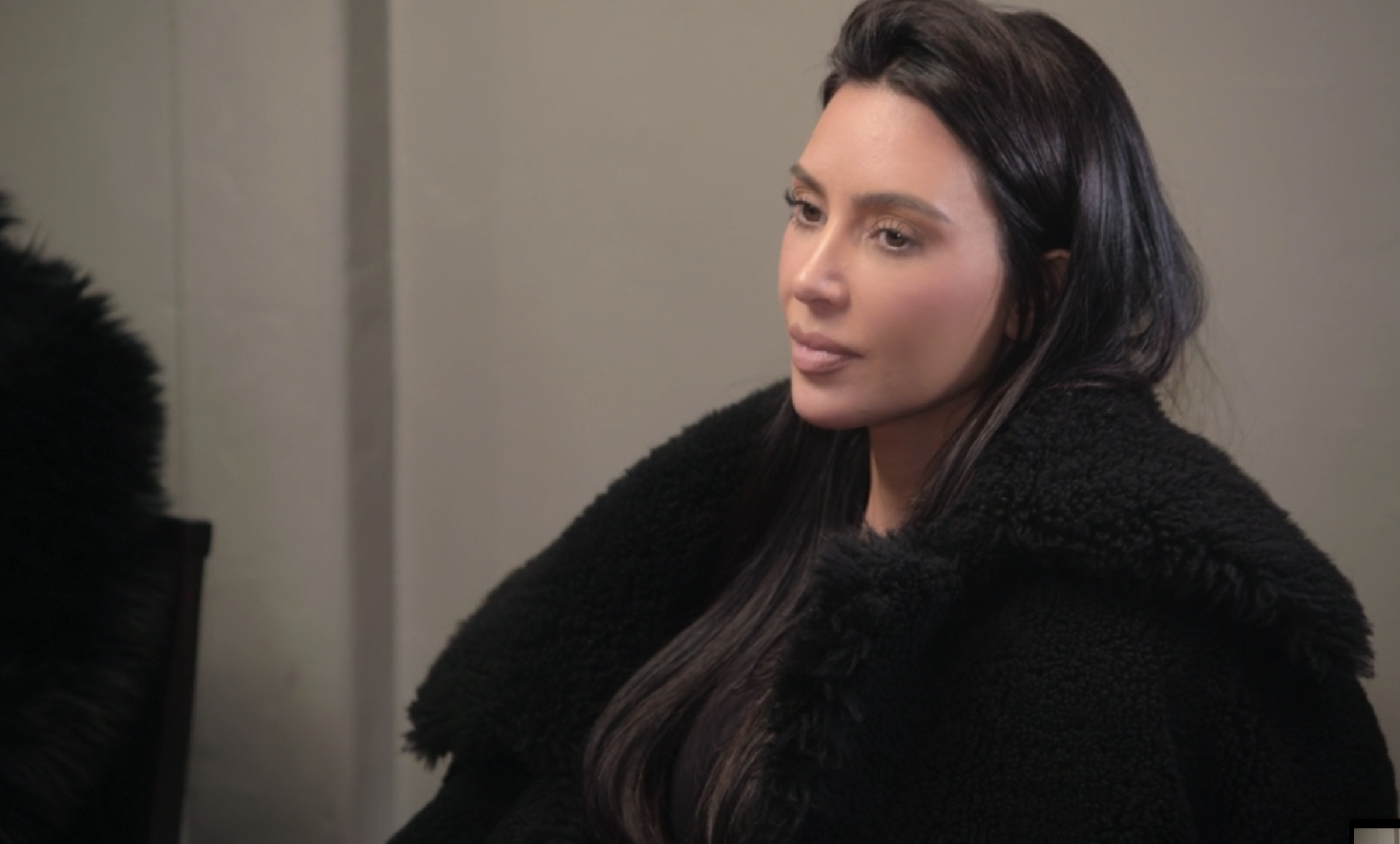 Kim Kardashian sits in a formal setting, wearing a black fur coat