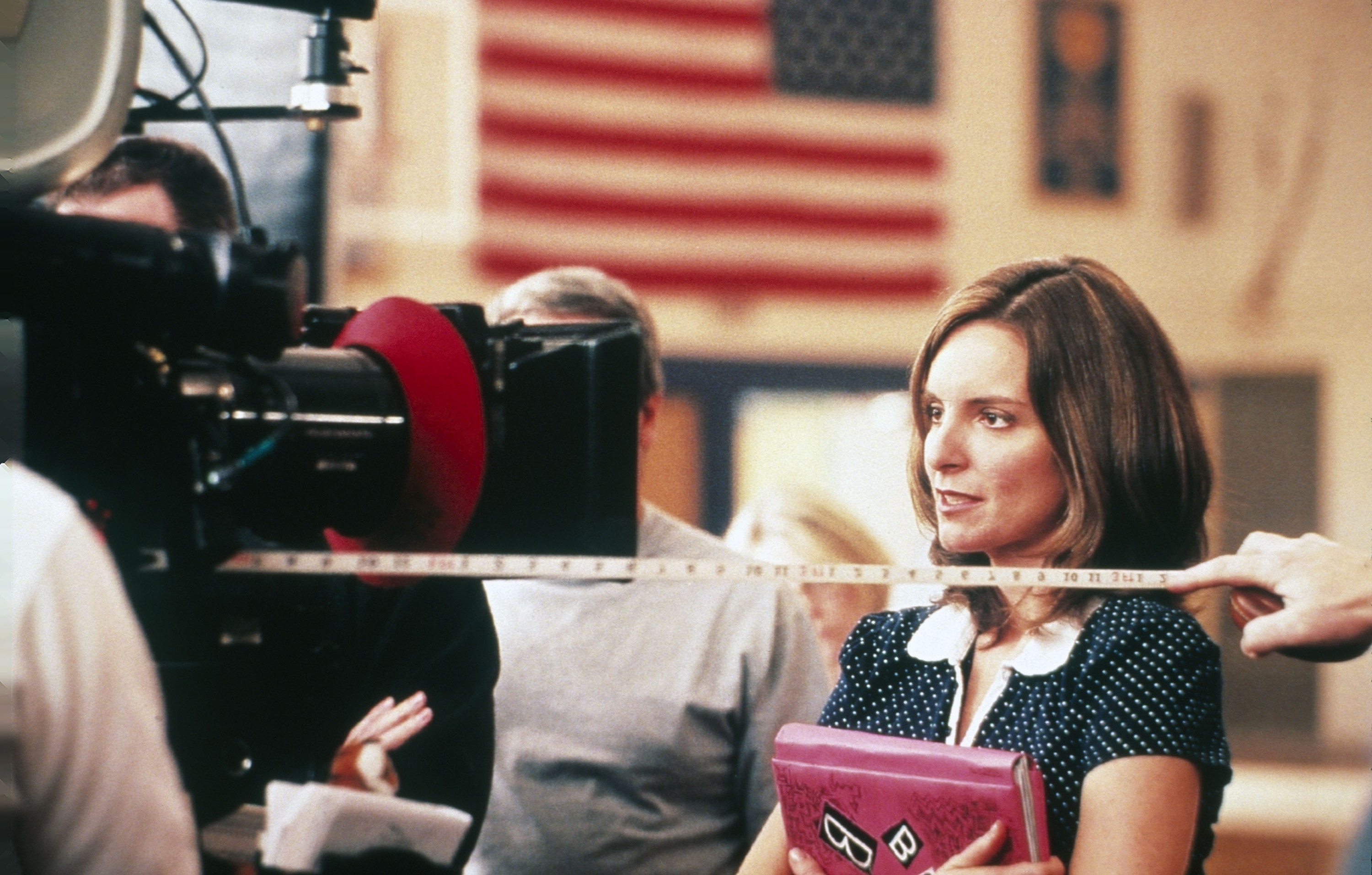 Tina Fey on a film set, holding a pink binder, with an American flag in the background. Crew members are adjusting equipment around her