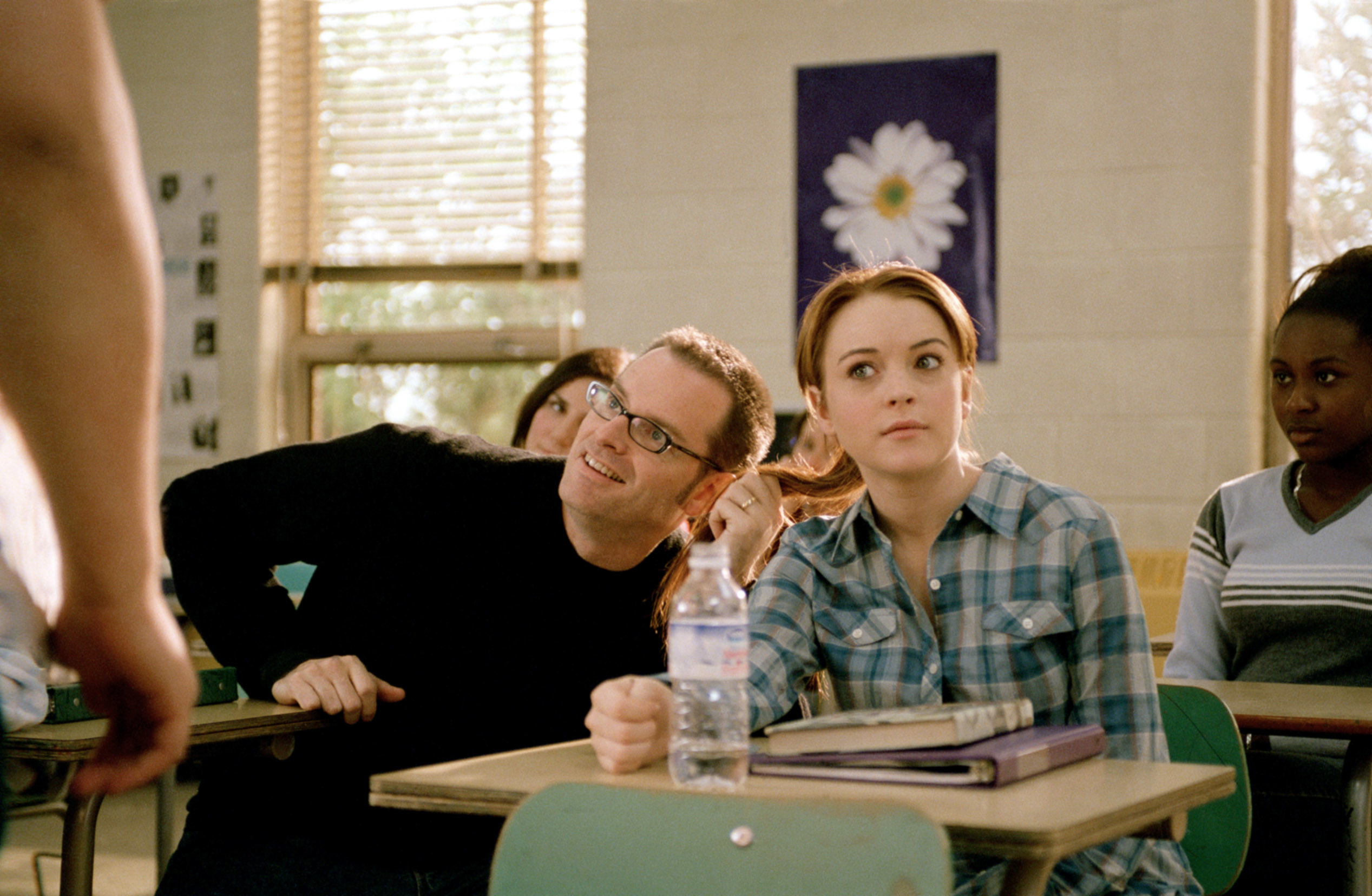 Lindsay Lohan in a classroom scene from a movie, looking surprised with a water bottle and notebooks on the desk