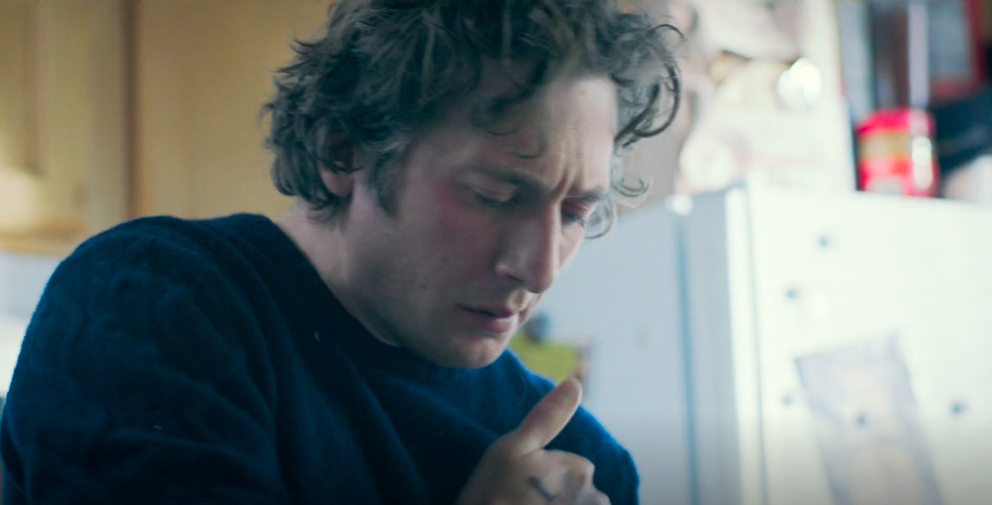 A man with curly hair, wearing a sweater, appears to be in thought or concern, in a kitchen setting. There are cabinets and a refrigerator in the background