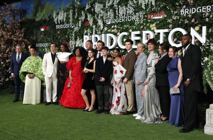 Cast of &quot;Bridgerton&quot; posed in front of a floral backdrop, wearing various elegant outfits on the red carpet