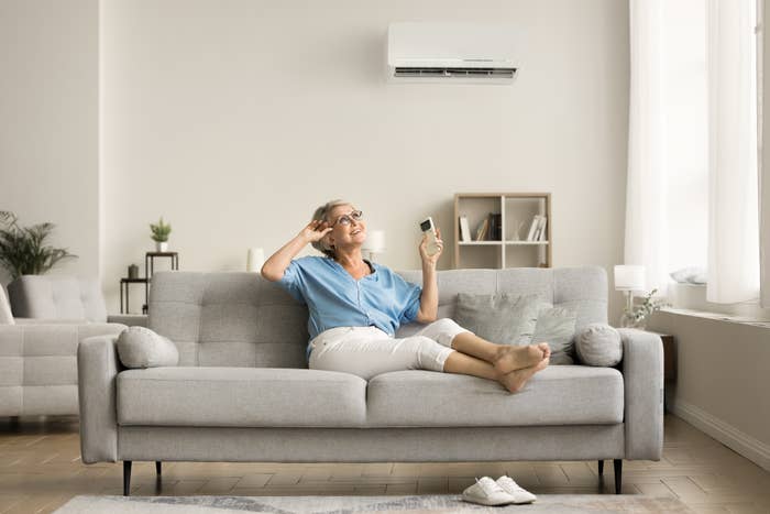 Older woman relaxing on a sofa with her feet up, holding a remote control, enjoying the air conditioning in a modern, well-lit living room