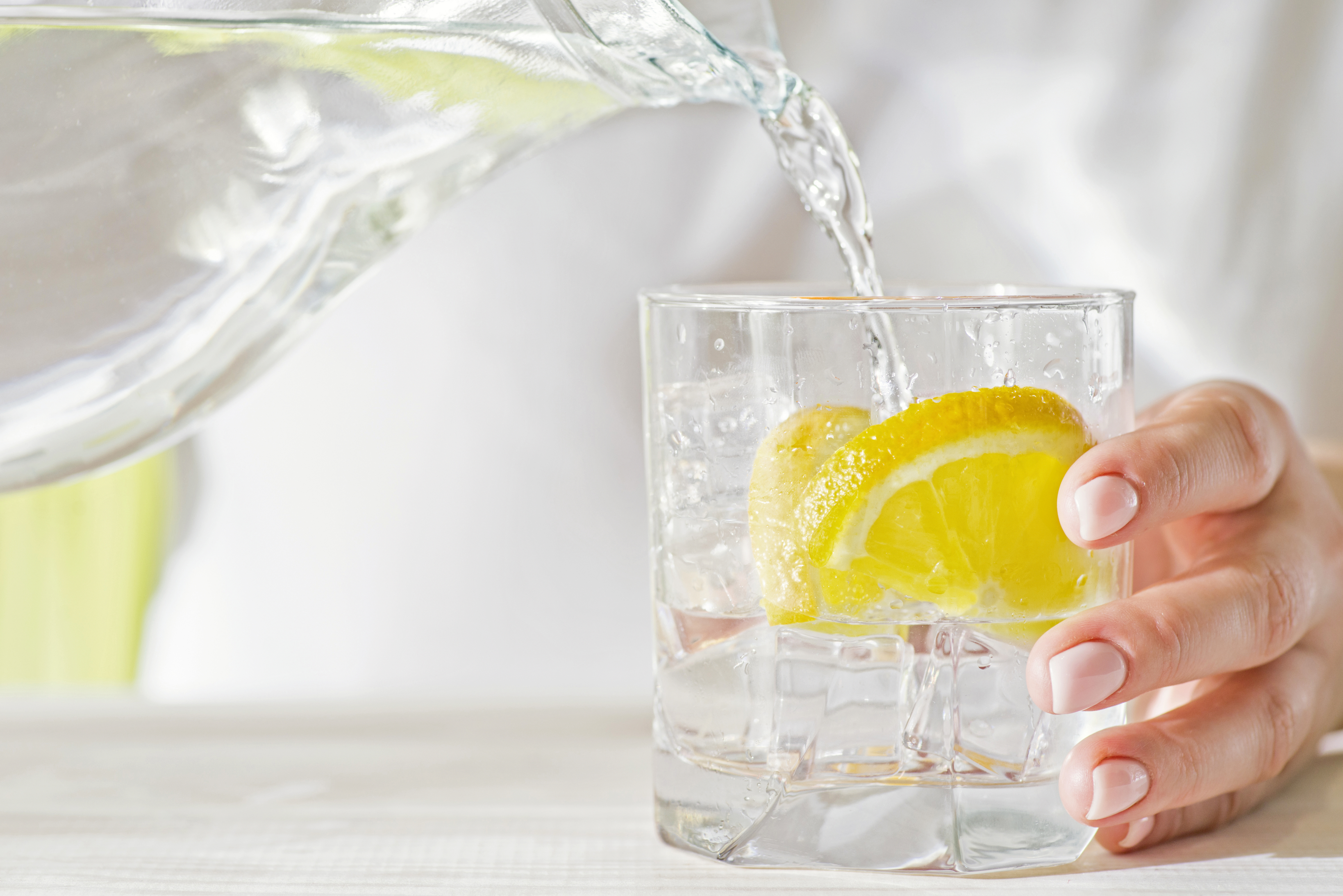 A hand pours water from a pitcher into a glass containing lemon slices