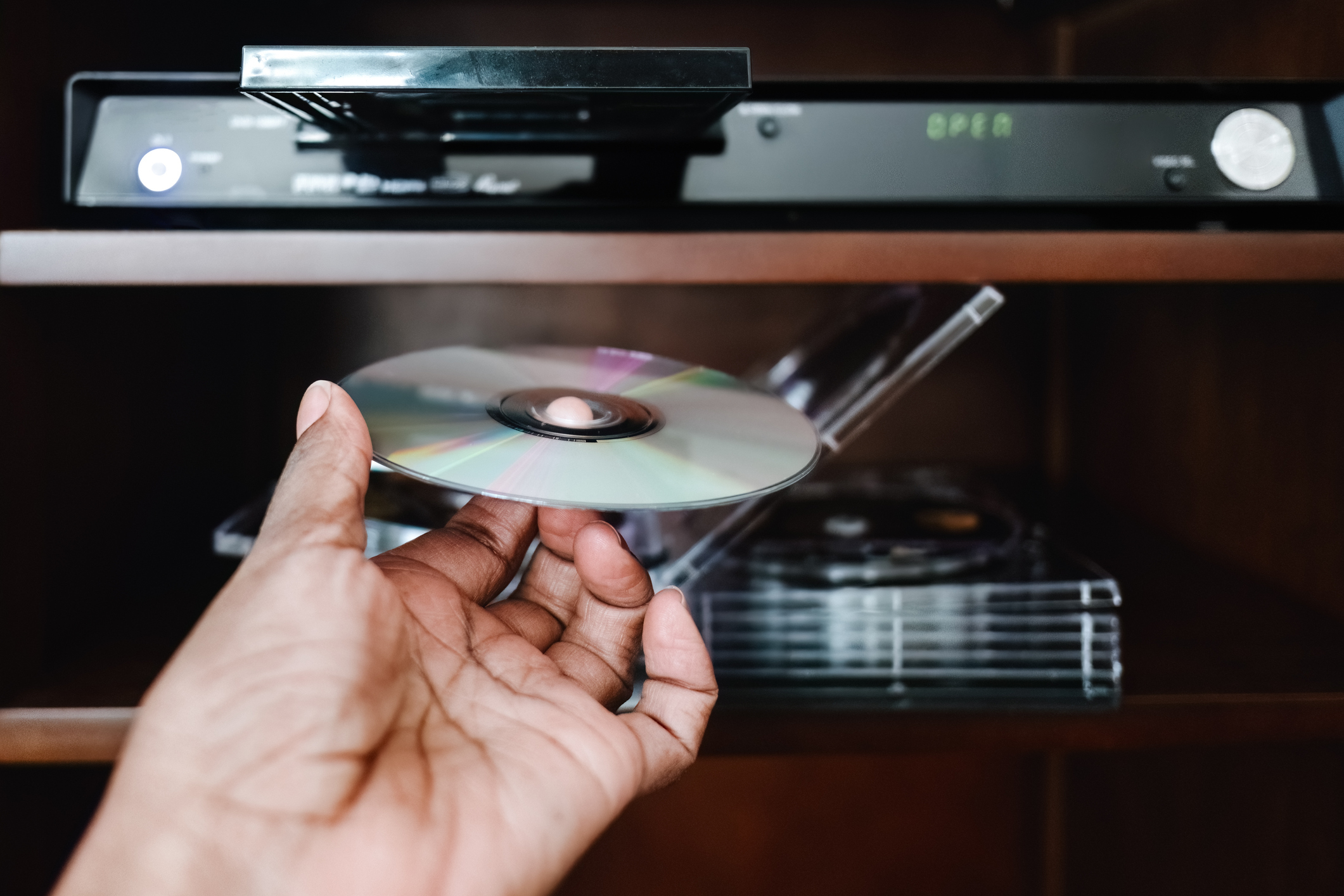 A hand inserting a DVD into a DVD player shelf, with several other DVDs visible. The DVD player is turned on