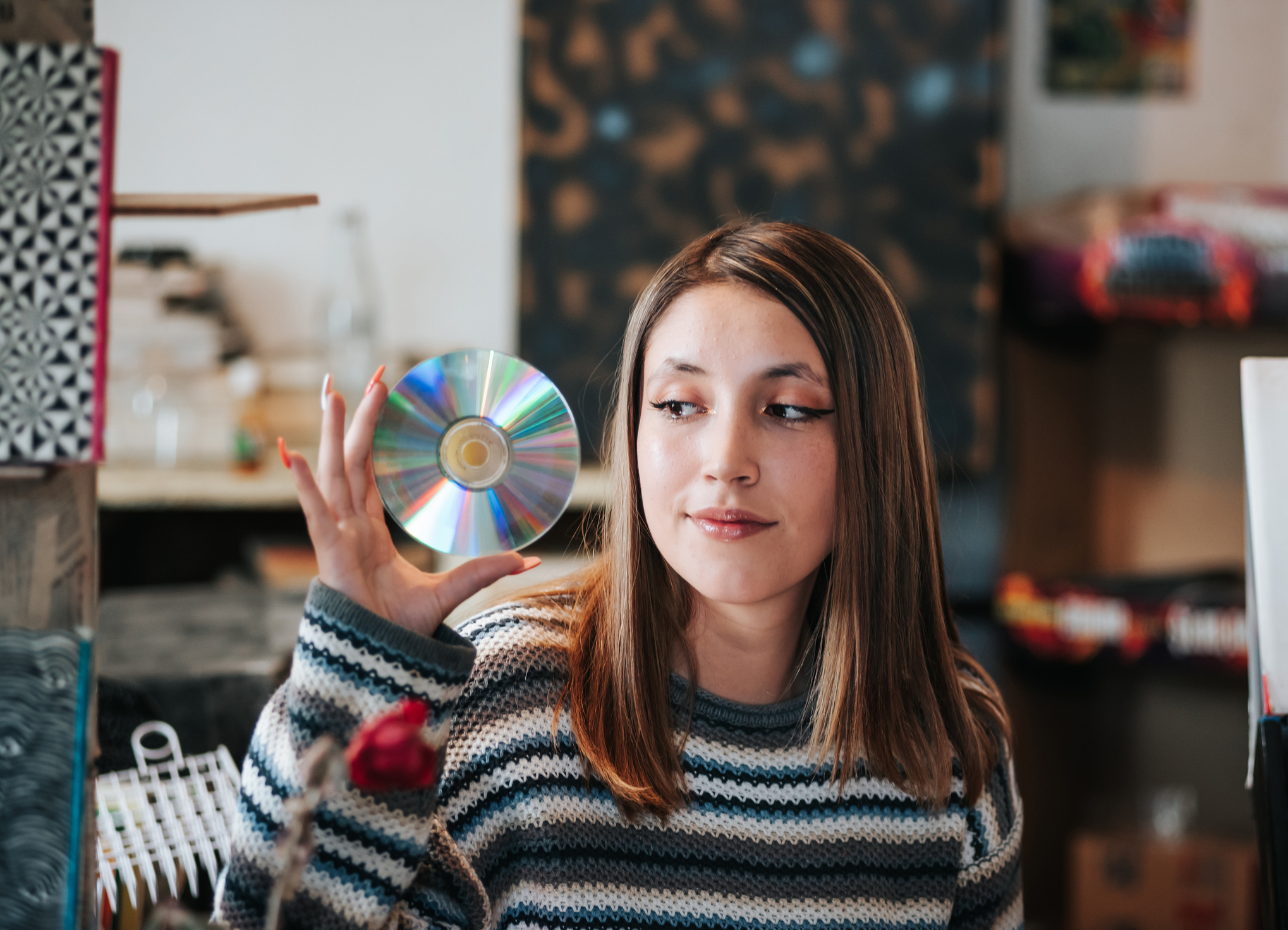 A woman in a striped sweater holds up a CD while looking at it, with a thoughtful expression