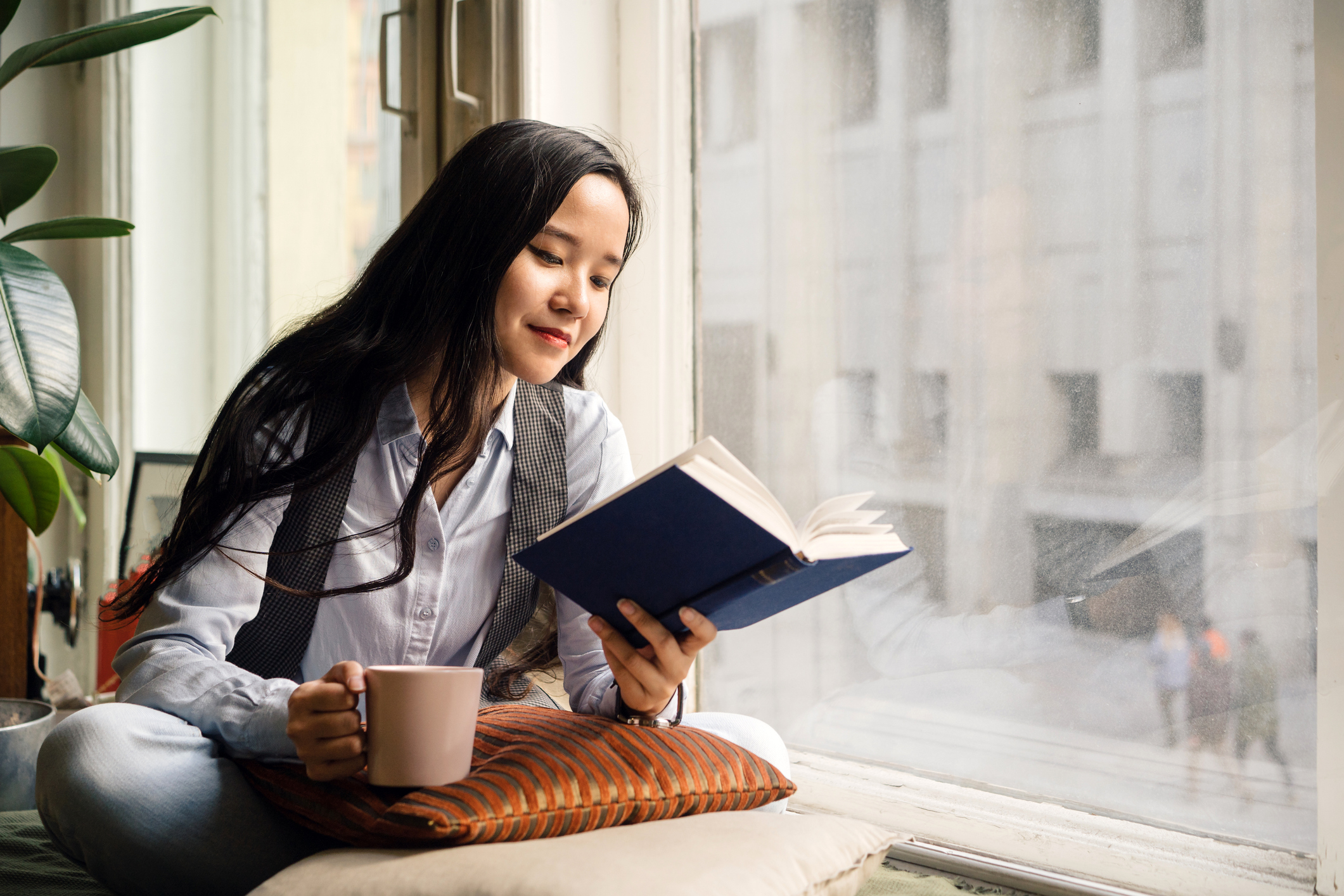 A woman with long hair reads a book by a window while holding a mug