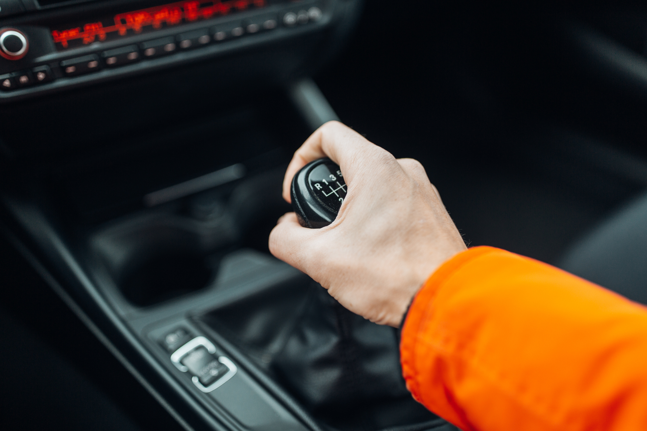 A hand wearing an orange sleeve is gripping the gear shift of a manual transmission car. The car&#x27;s interior is partially visible