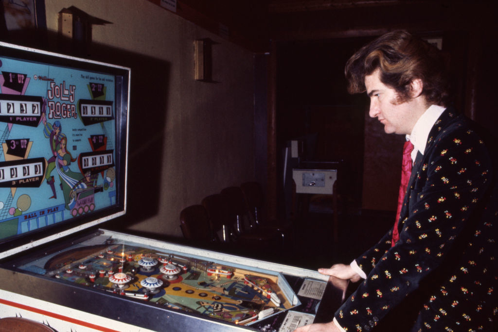 A man in a stylish suit with a patterned jacket and pink tie playing a vintage pinball machine named &quot;Jolly Poker&quot; in a dimly lit arcade