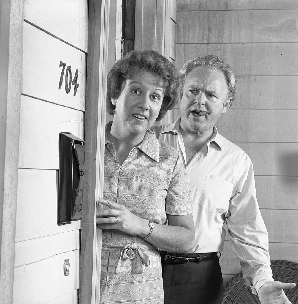 Jean Stapleton and Carroll O&#x27;Connor standing at the door labeled 704, appearing surprised. Jean Stapleton is wearing a patterned dress, while Carroll O&#x27;Connor is in a light shirt and dark pants