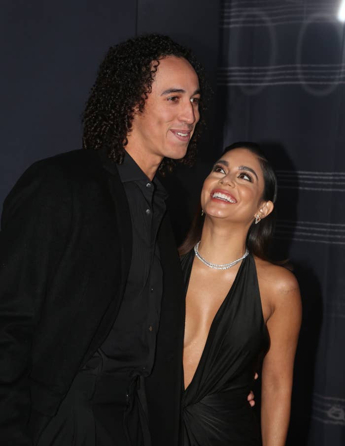 Cole and Vanessa Hudgens smile while posing on a red carpet. She wears a plunging halter dress and a sparkling necklace