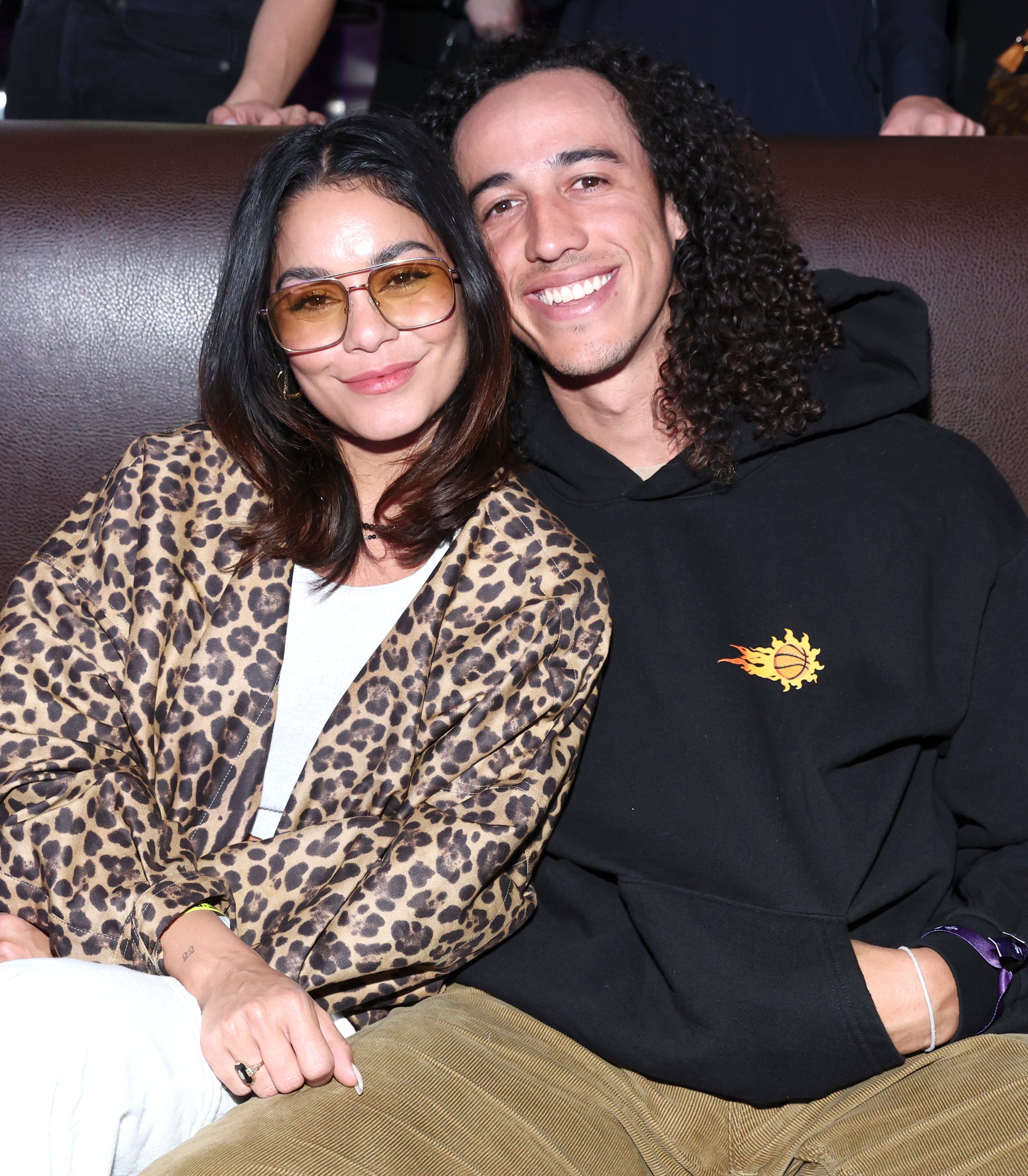 Vanessa Hudgens in a leopard-print coat and Cole Tucker in a black hoodie, sit together smiling on a couch