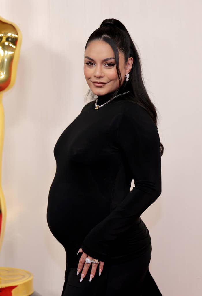 A pregnant Vanessa Hudgens in a sleek, long-sleeve dress, standing with a hand on her hip at an event. She has styled her hair in a high ponytail