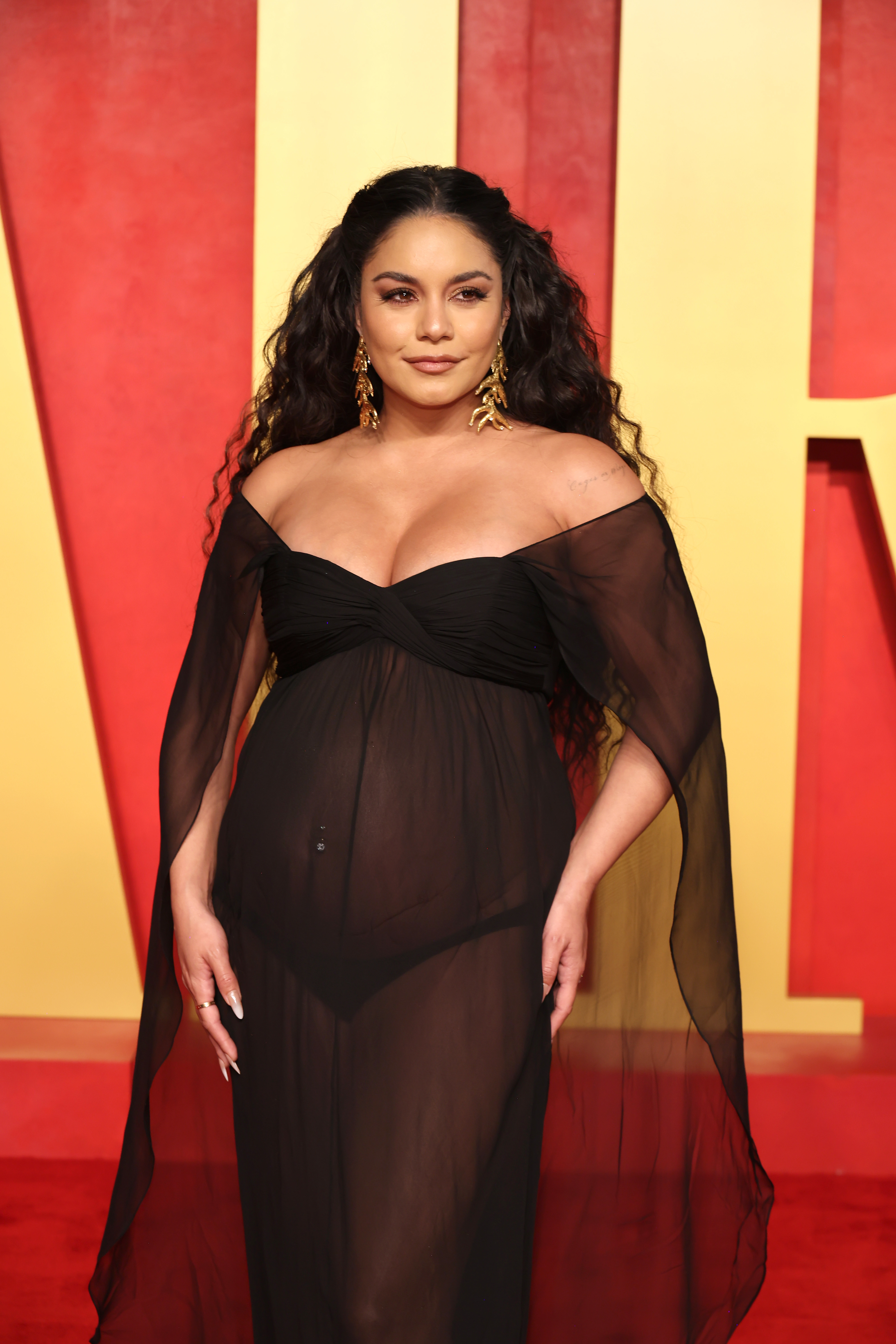 Vanessa Hudgens on a red carpet wearing a off-the-shoulder, sheer gown with gold earrings