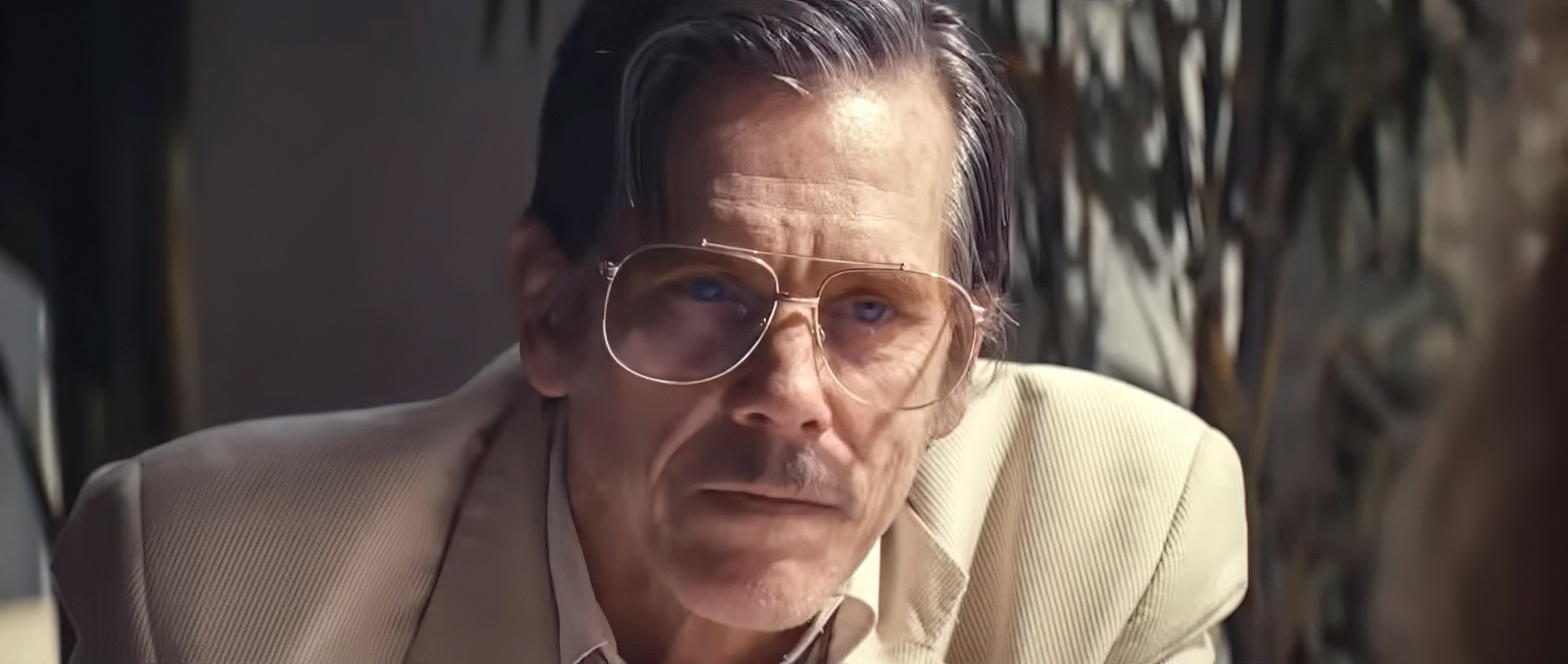 Kevin Bacon wears glasses and a light-colored blazer, looking intently at someone off-camera
