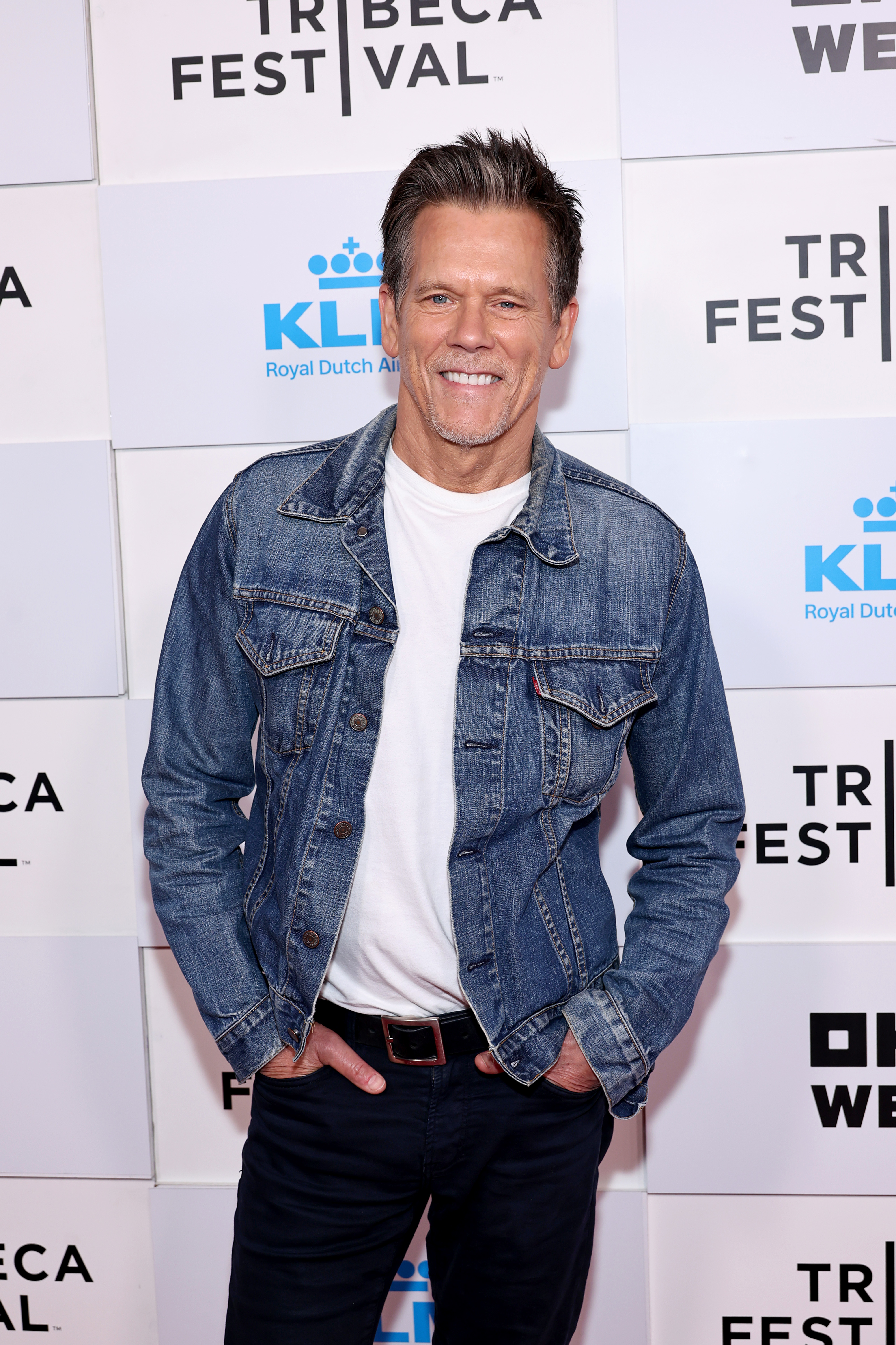 Kevin Bacon poses at the Tribeca Festival, wearing a casual denim jacket over a white shirt and black pants. He smiles with hands in his pockets