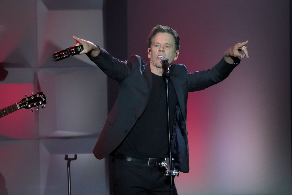 Kevin Bacon performs on stage, singing into a microphone with arms outstretched, dressed in a dark suit and black shirt