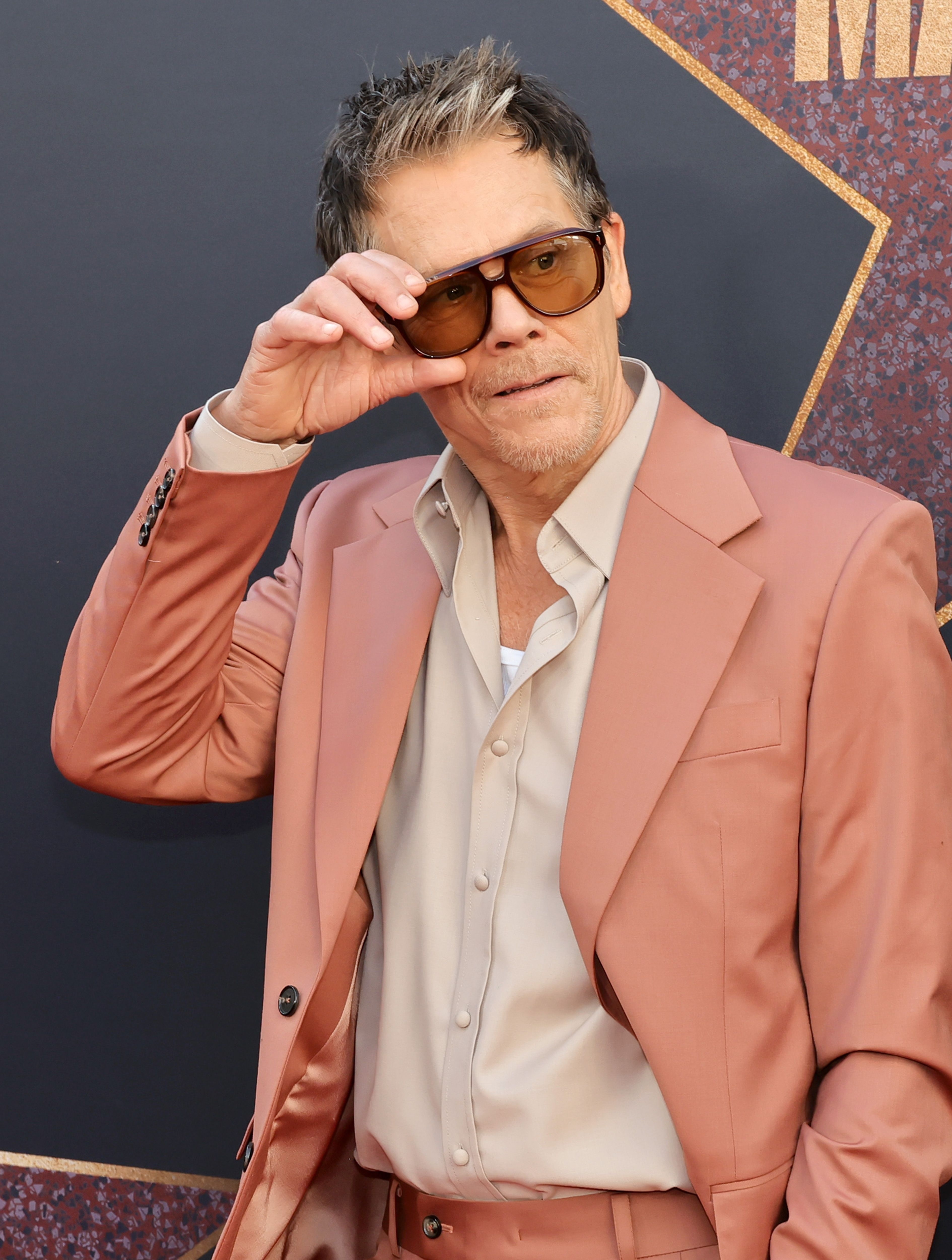 Kevin Bacon on the red carpet, wearing a light beige shirt and a matching suit, with sunglasses