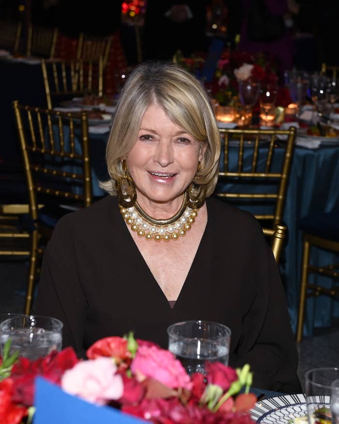 Martha Stewart at a formal dinner event, wearing a black dress and a statement pearl necklace, seated among elegant table settings and floral centerpieces