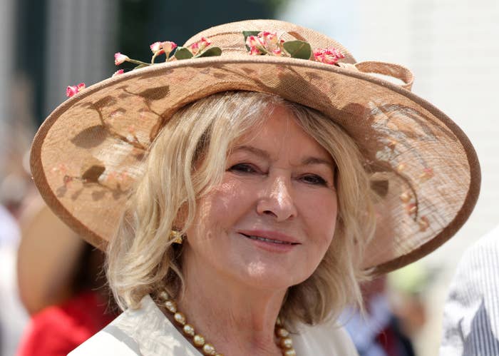 Martha Stewart wearing a floral-decorated wide-brim hat and pearl necklace, smiling at an outdoor event