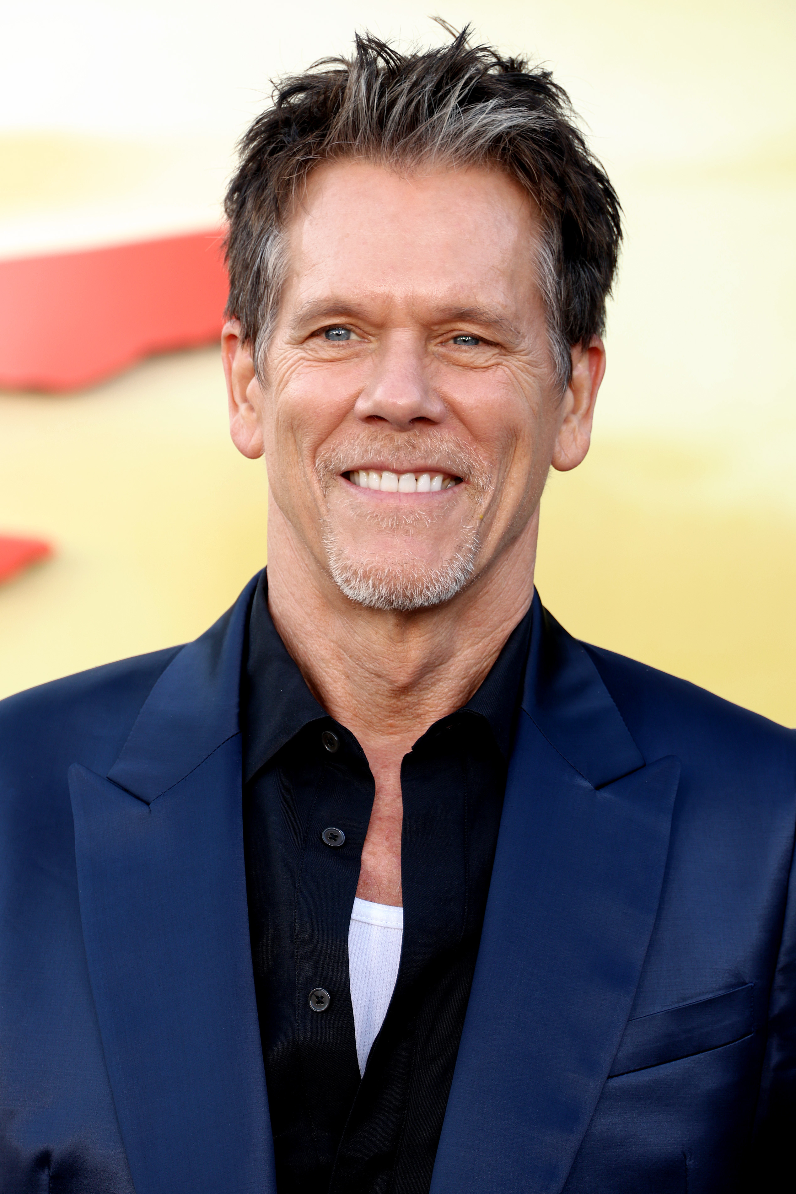 Kevin Bacon smiles on the red carpet, wearing a dark button-down shirt under a blazer