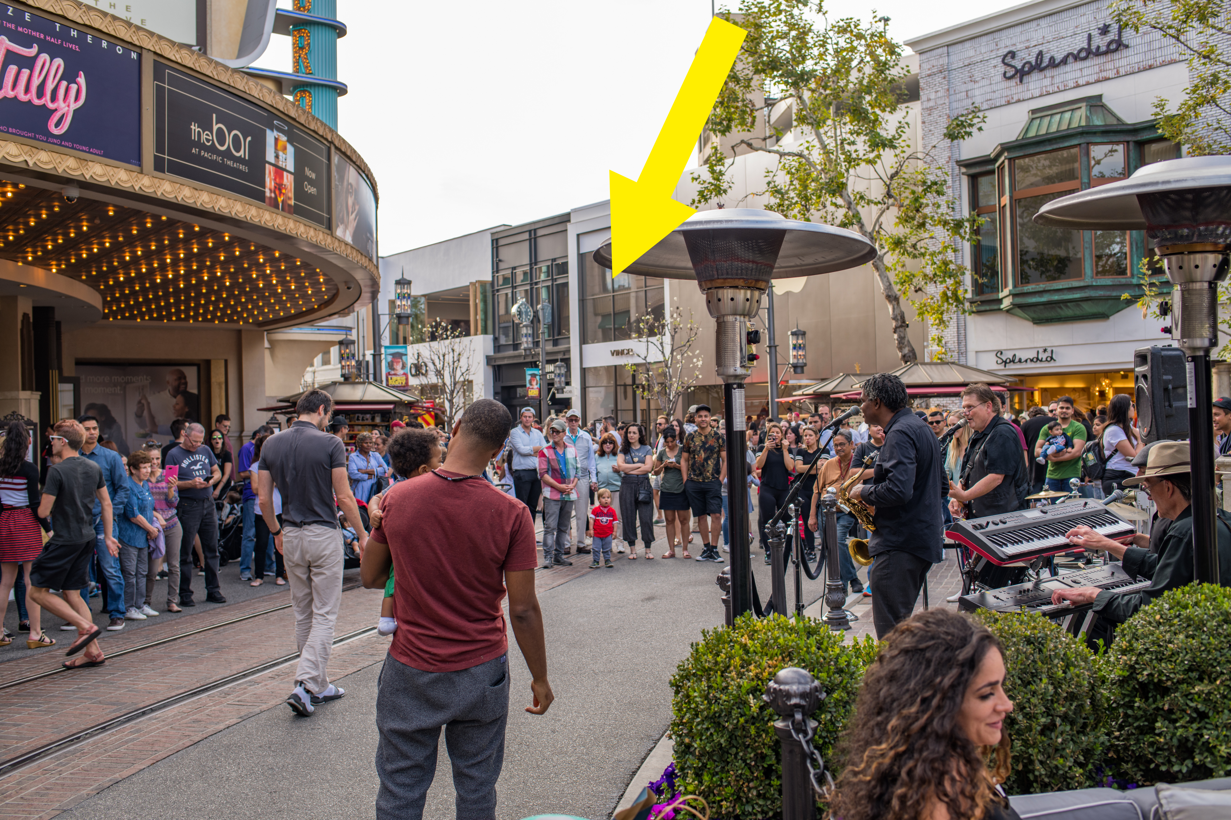 A crowd gathers around street musicians performing outside a theater. Restaurant signs and various shops are visible in the background