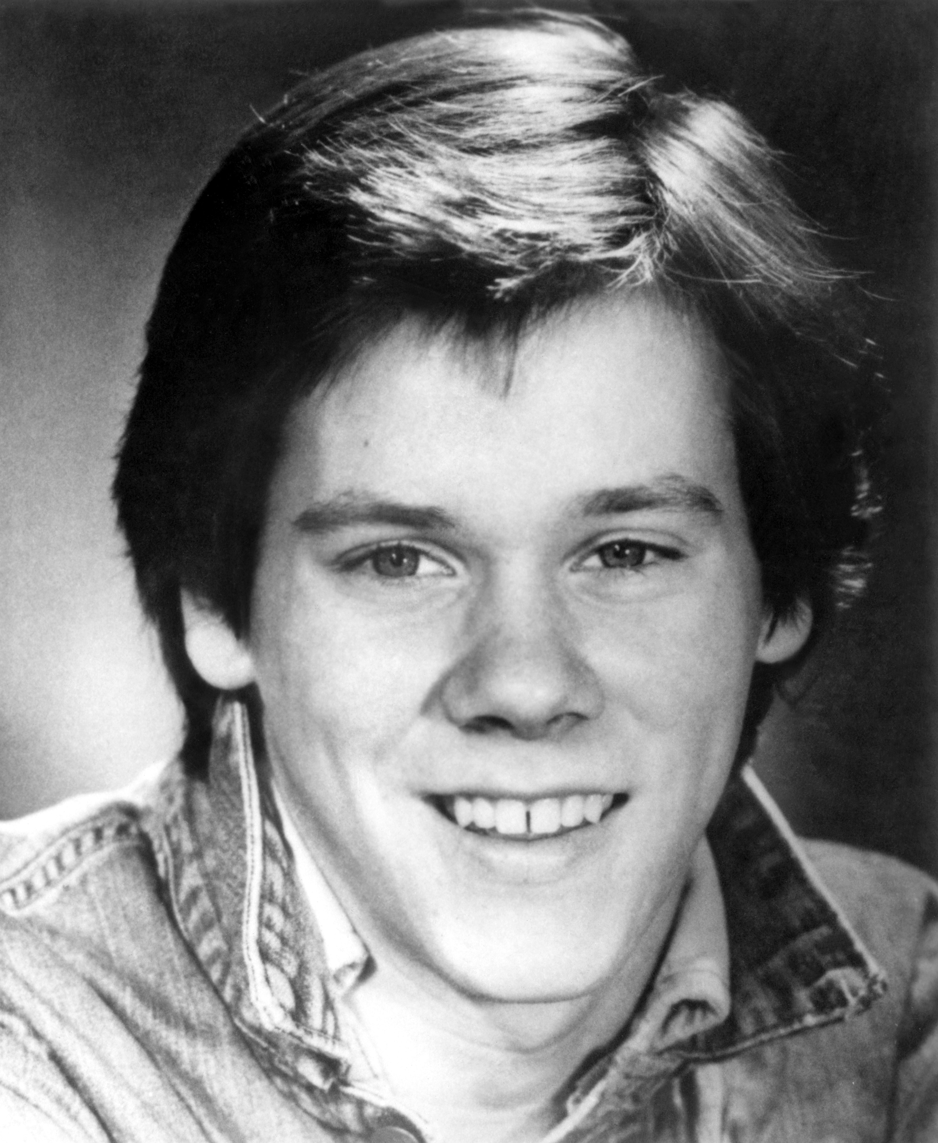 Young Kevin Bacon smiling with medium-length hair, wearing a casual collared shirt and denim jacket